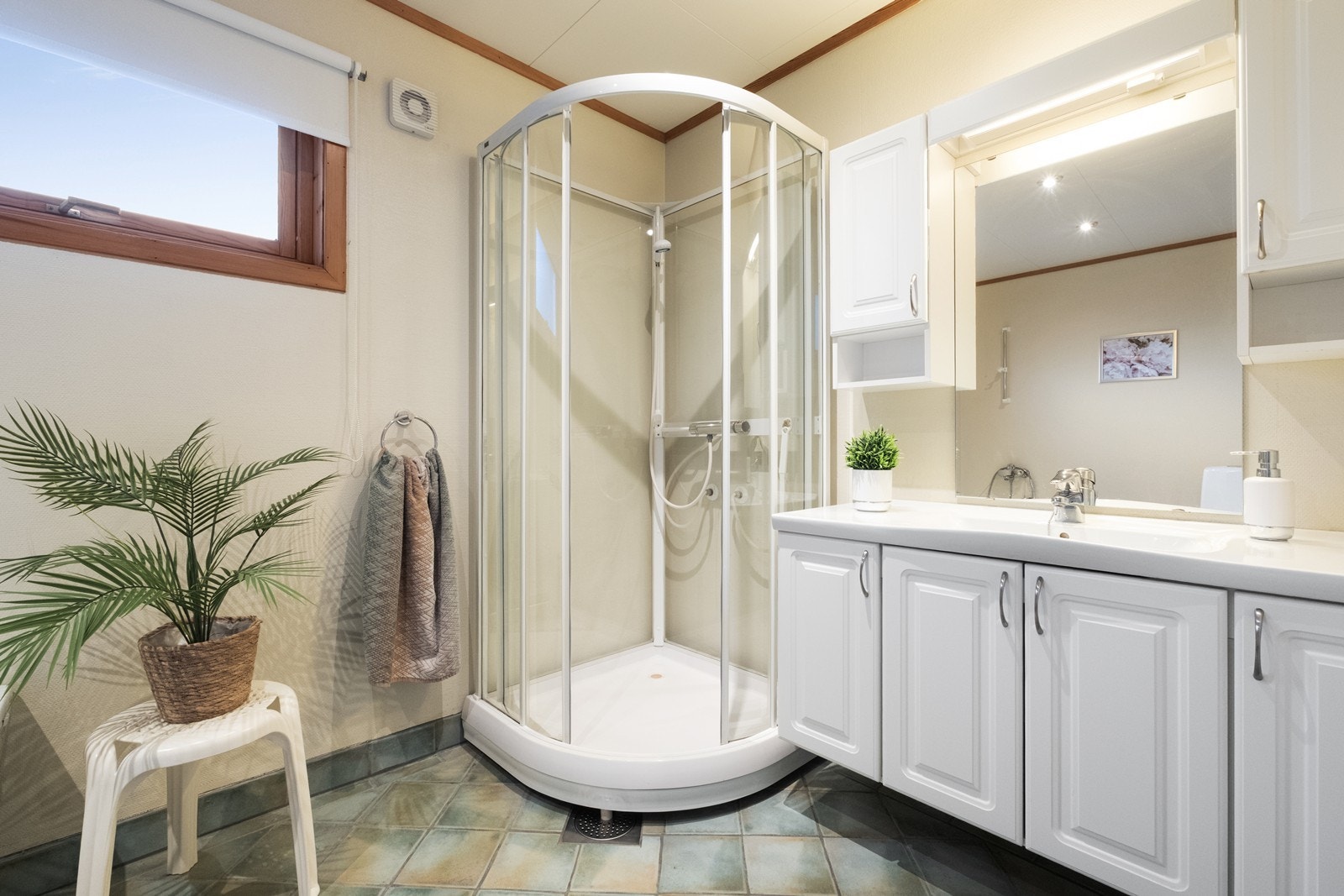 Badet har flislagt gulv og er innredet med dusjkabinett og hvit baderomsinnredning med både over- og underskap og heldekkende servant. Galleribilde