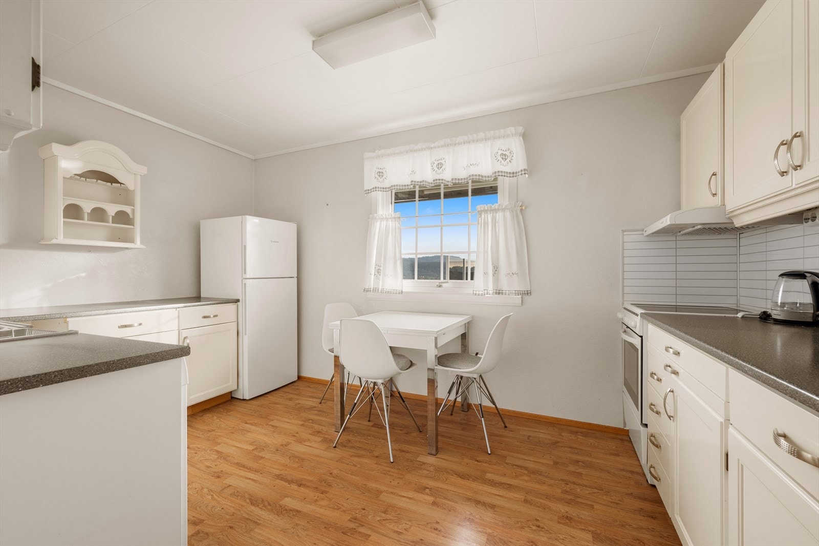 Kjøkkenet har en innredning med lyse, profilerte fronter, laminert benkeplate og flisplate på vegg mellom benkeplate og overskap. Galleribilde