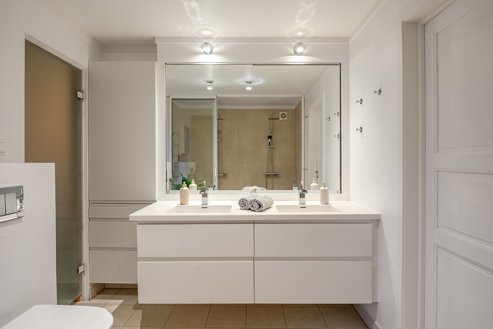 Moderne baderomsinnredning med skuffer og heldekkende dobbel servant med fossefall-armaturer, stort speil med belysning over. Ekstra oppbevaringsplass i høy vegghengt innredning ved siden av. Galleribilde