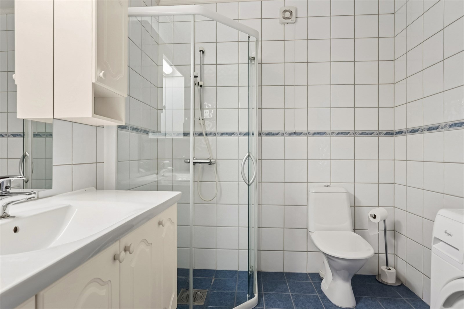 Badet har fliser på gulvet og på veggene, og himlingsplater. Rommet er innredet med servant med under- og overskap, speil med tilhørende belysning, dusvegger og gulvmontert toalett. Galleribilde