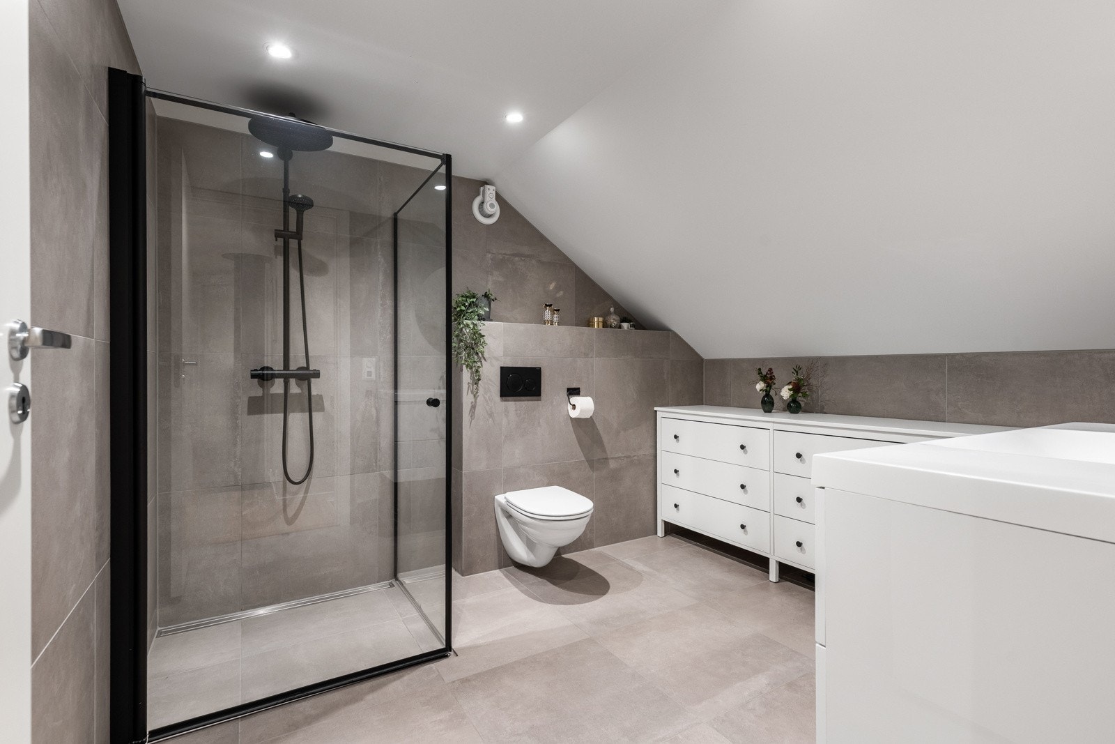 Hovedbadet i andre etasje viderefører den moderne og stilrene utformingen fra første etasje, med flislagte overflater, servant med speil, vegghengt toalett og dusjhjørne med svingbare glassdører Galleribilde