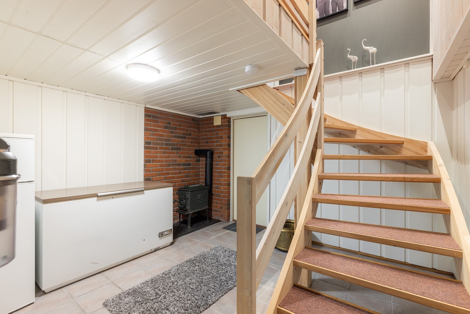 Fra hovedetasjen er det åpen trapp ned til sokkeletasjen hvor det er gang, bad/vaskerom, soverom og boder. Gangen har flislagt gulv med varmekabler og det er montert vedovn. Galleribilde