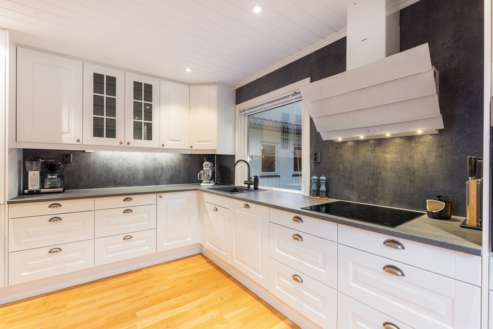 Kjøkkenet har alle hvitevarer integrert: oppvaskmaskin, induksjonstopp og kjøl/fryseskap, samt stekeovn plassert i praktisk arbeidshøyde i høyskap. Over koketoppen er det en stilfull ventilator med utlufting via yttervegg. Vannfaste plater på vegg. Galleribilde