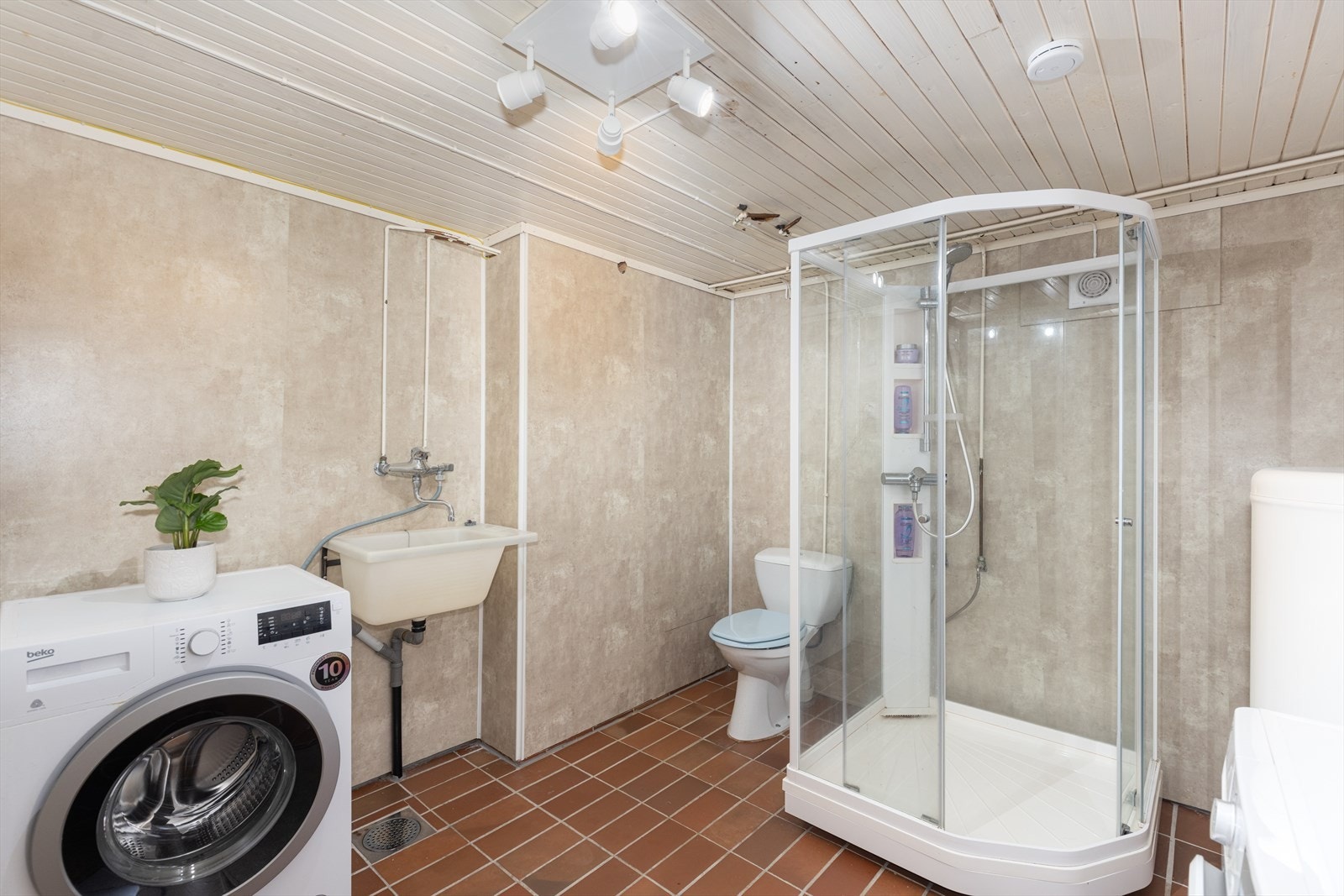 I sokkeletasjen er det et kombinert bad/vaskerom med keramiske fliser på gulvet og varmekabler. Her er det dusjkabinett, toalett, vaskekum og opplegg for vaskemaskin. Øvrige overflater er plater på vegg og malt panel i tak. Galleribilde