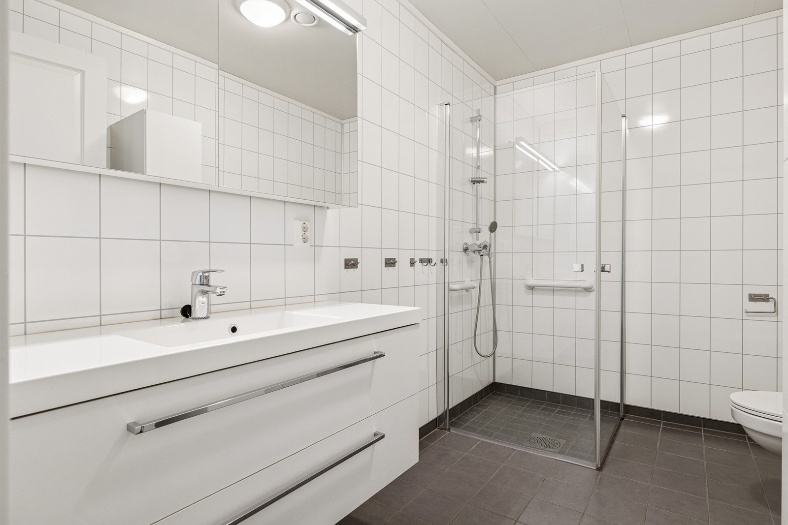 Moderne, lyst og funksjonelt bad. Veggene er kledd med store, hvite fliser som gir rommet et rent og tidløst uttrykk, mens det mørke grå gulvet skaper en fin kontrast og gir en følelse av eleganse. Galleribilde