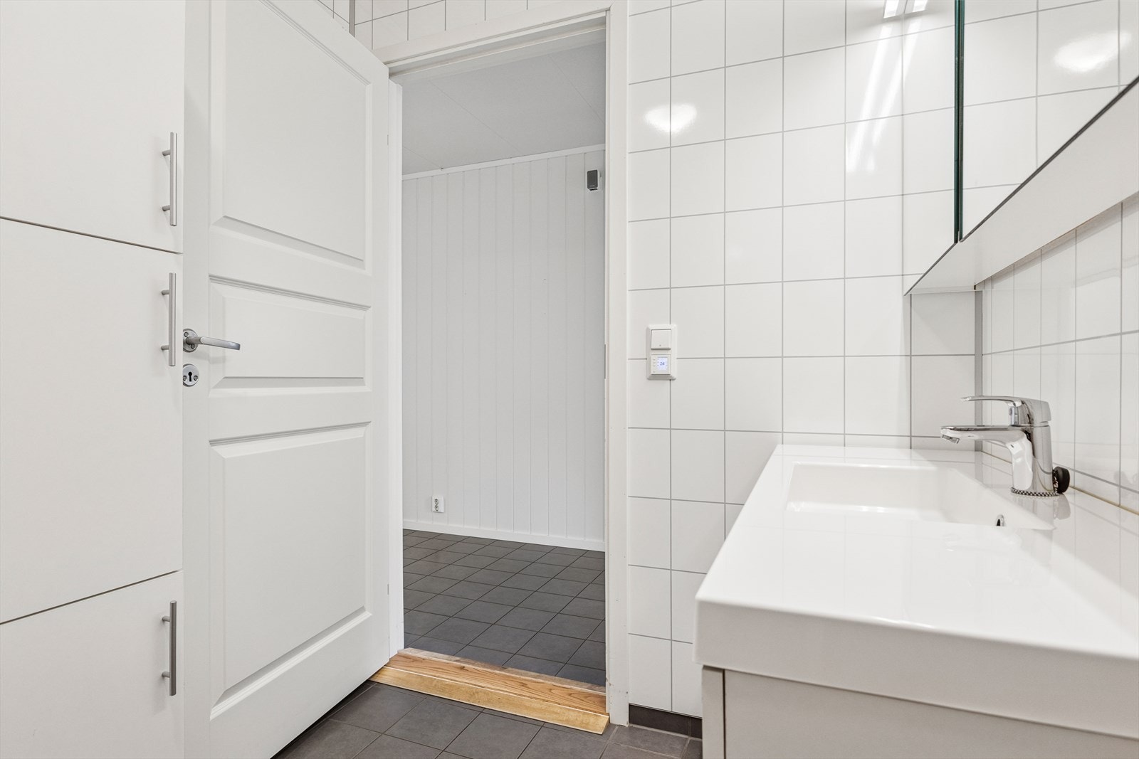 Baderomsinnredningen er stilren, med en hvit, minimalistisk servant med god skapplass under og to skuffer med elegante, krommede håndtak. Over servanten er det et stort speil med integrert belysning som gir godt lys og åpner opp rommet ytterligere. Galleribilde