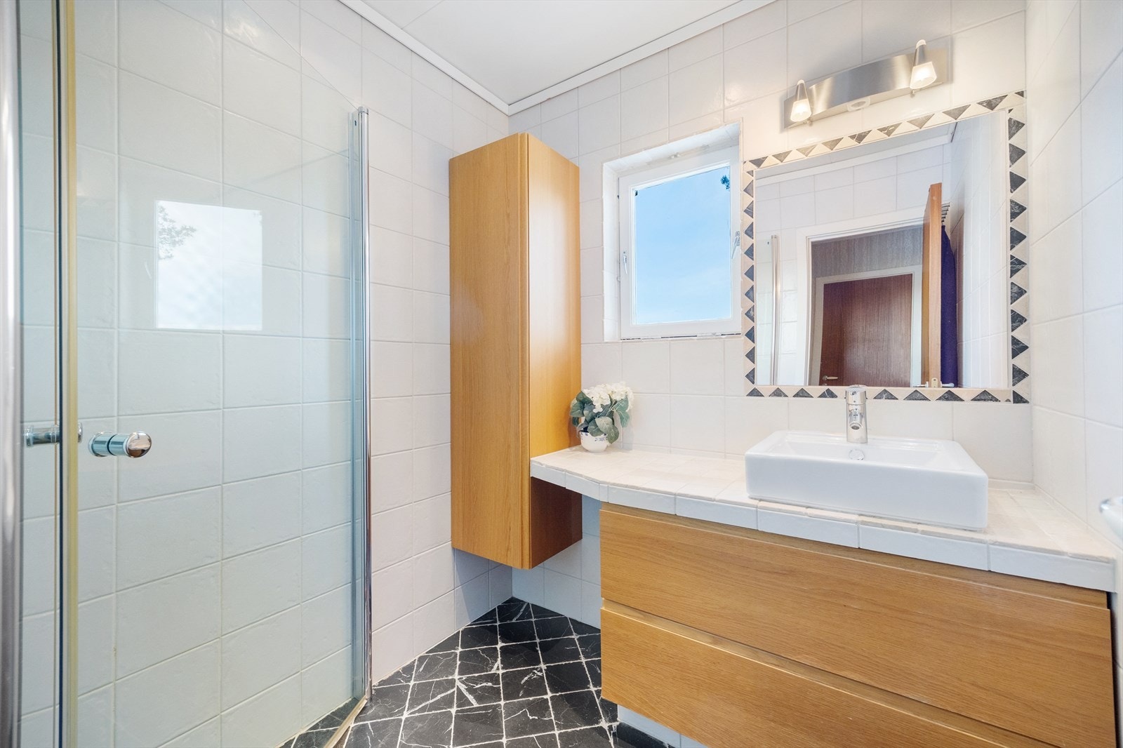 Badet er helfliset med marmorfliser og elektriske varmekabler i gulvet. Rommet er innredet med innfellbart dusjhjørne i glass, fliselagt benkeplate med servant og innfliset speil, samt vegghengt skap i eik Galleribilde