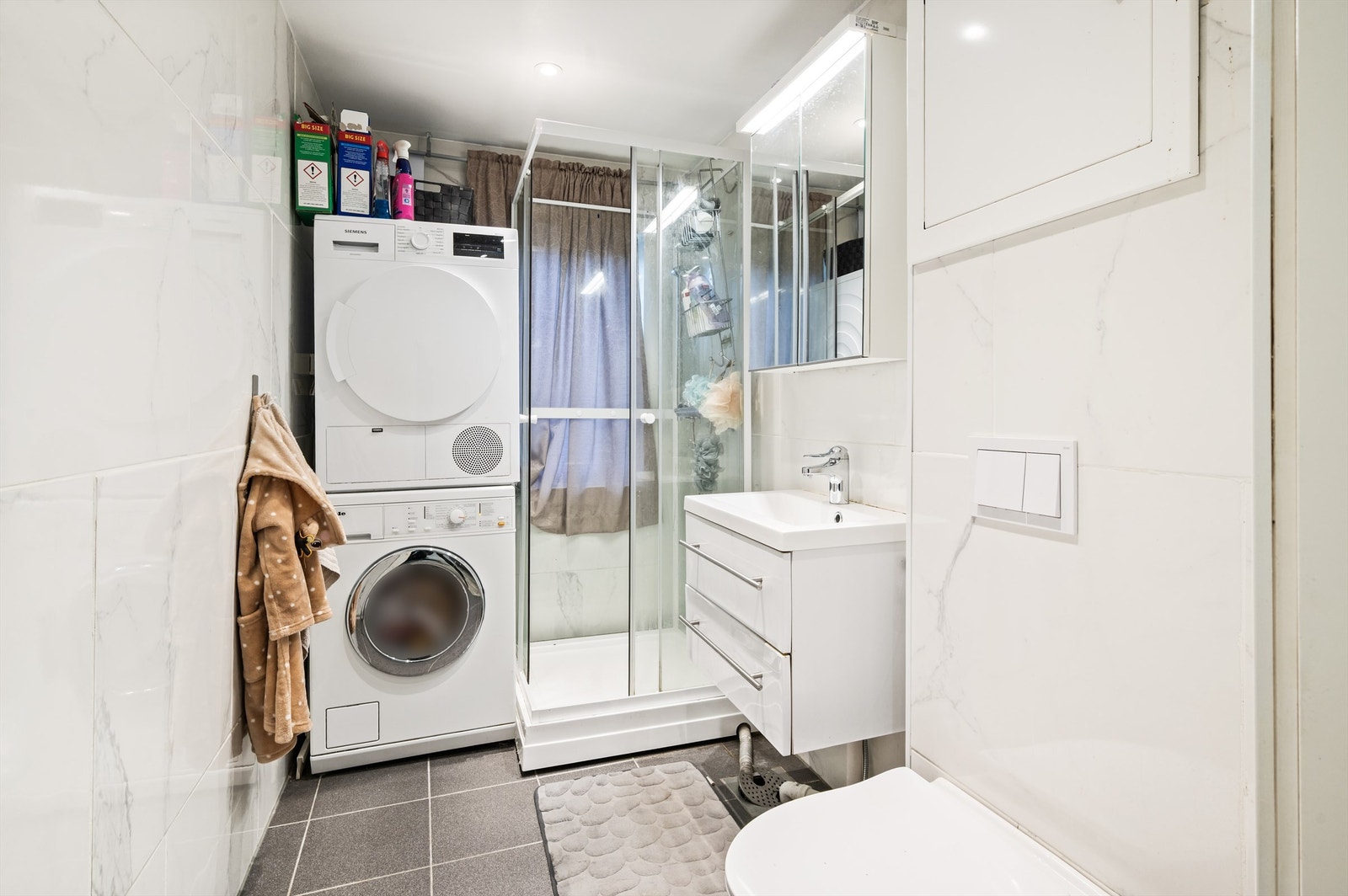 Badet er flislagt på gulv og vegger med varme i gulv. Innredningen består av blant annet vegghengt servantskap og toalett med innebygget sisterne. Galleribilde