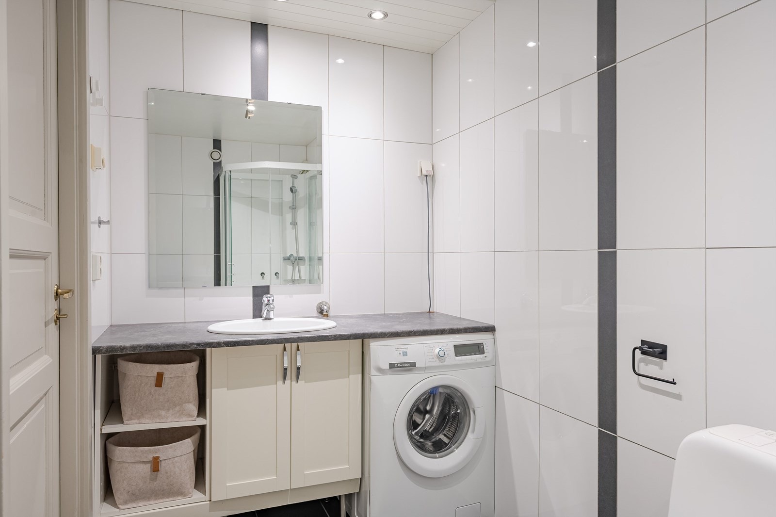 Flislagt baderom med servant på innredning, speil, hyller for oppbevaring og opplegg for vaskemaskin. Galleribilde