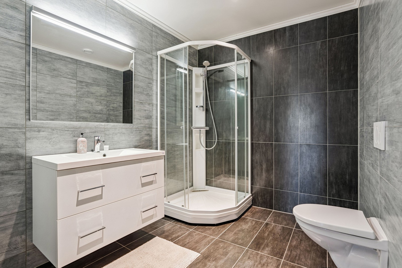 Gulvet er flislagt og utstyrt med gulvvarme. Baderomsinnredning fra Linnbad, med heldekkende servant og glatte fronter. På veggen er det speil med integrert belysning. Vegghengt toalett og dusjkabinett Galleribilde