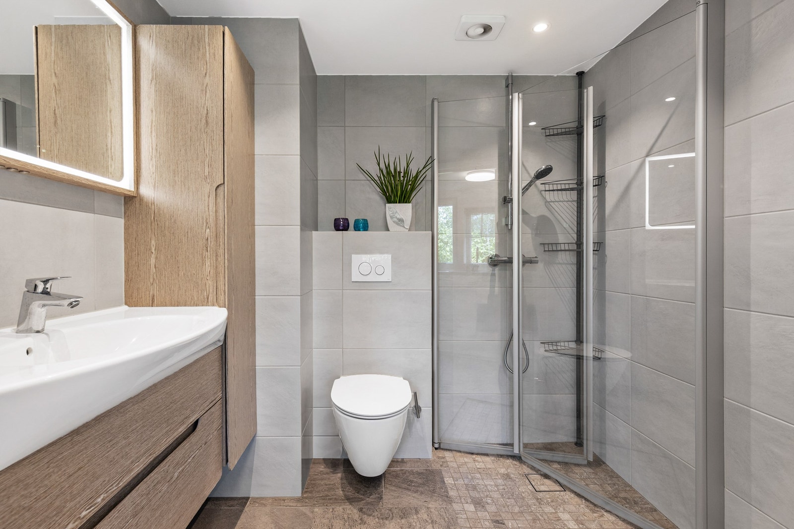 Badet er pent flislagt på gulv og vegger med elektrisk varme i gulvet. Badet er innredet med vegghengt klosett, servant nedfelt i innredning, speil med lys og dusjhjørne med svingbare glassdører. Galleribilde