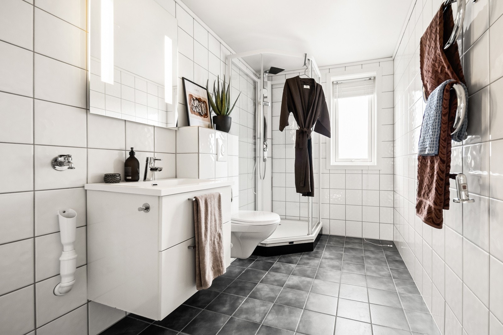 Flislagt baderom innredet med dusjkabinett, servant med skuffeseksjon, vegghengt klosett og opplegg for vaskemaskin Galleribilde