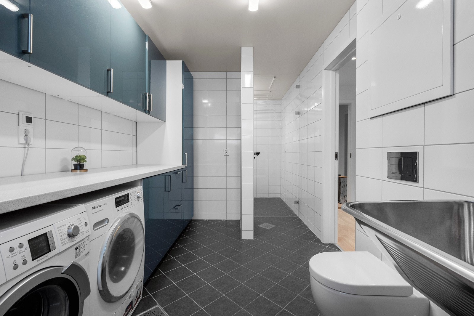 En veggmontert utslagsvask og et vegghengt toalett sikrer funksjonalitet, mens innredningen er tilrettelagt med opplegg for både vaskemaskin og tørketrommel
