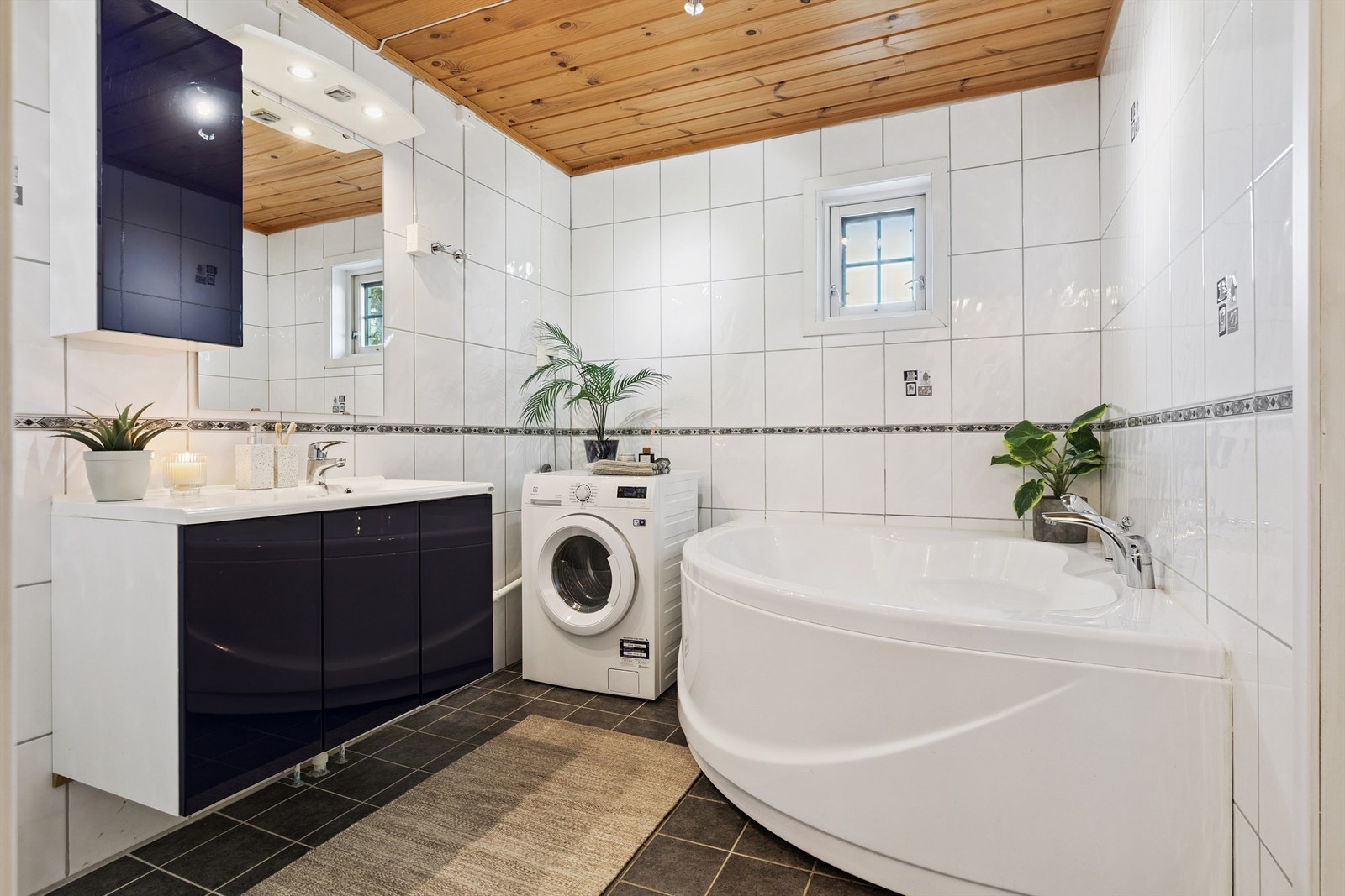 Bad i del 2: Badet har flislagt gulv, flislagte vegger og en himling med trepanel og belysning. Vegghengt speil, lys og overskap er også en del av interiøret. Hjørnebadekar. Galleribilde