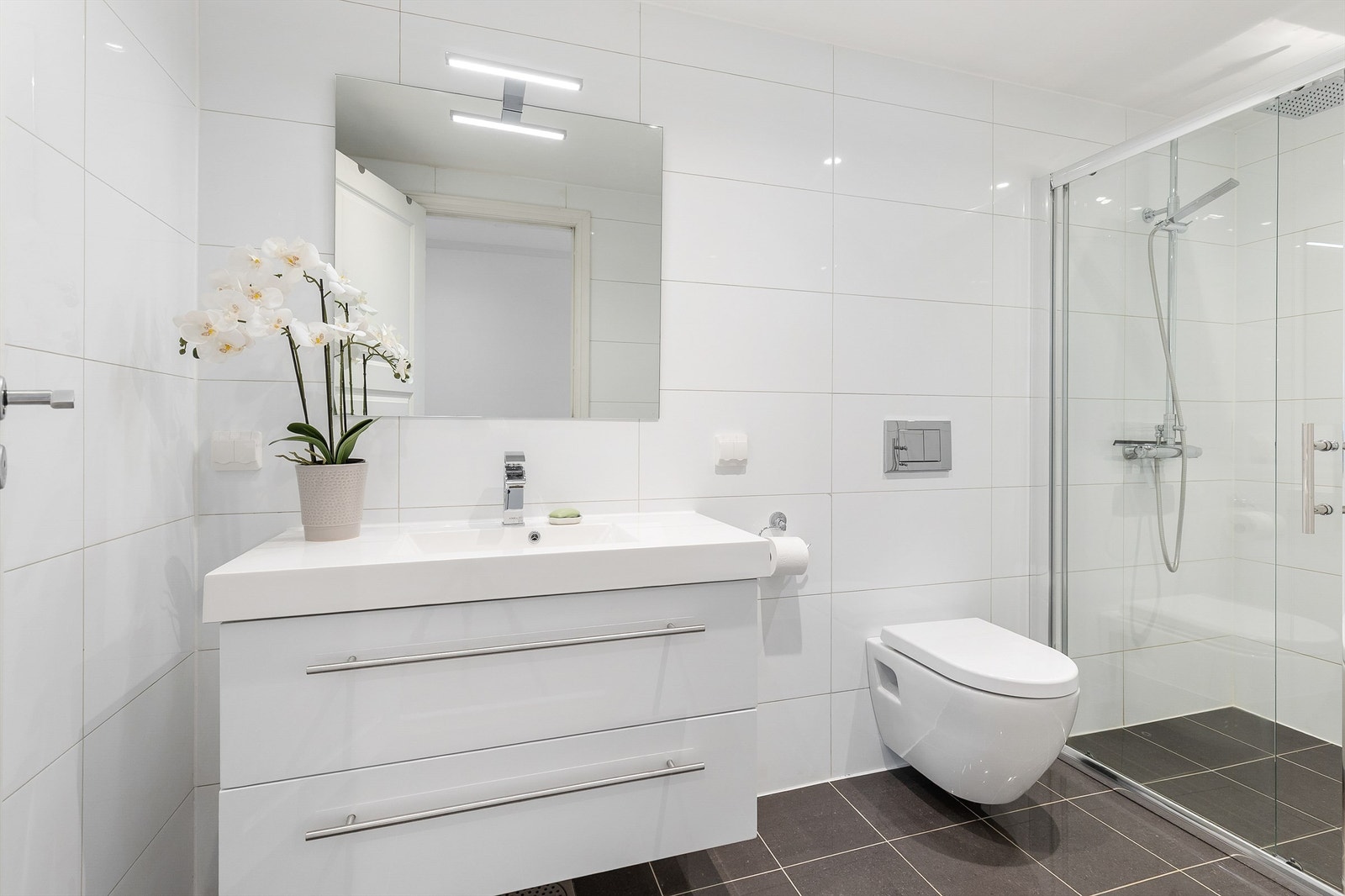 Badet i 1. etasje er pent flislagt på gulv og vegger, og har elektrisk gulvvarme som gir god komfort. Rommet har vegghengt toalett, servant nedfelt i innredning, speil med belysning og dusjhjørne med skyvedører i glass. Galleribilde