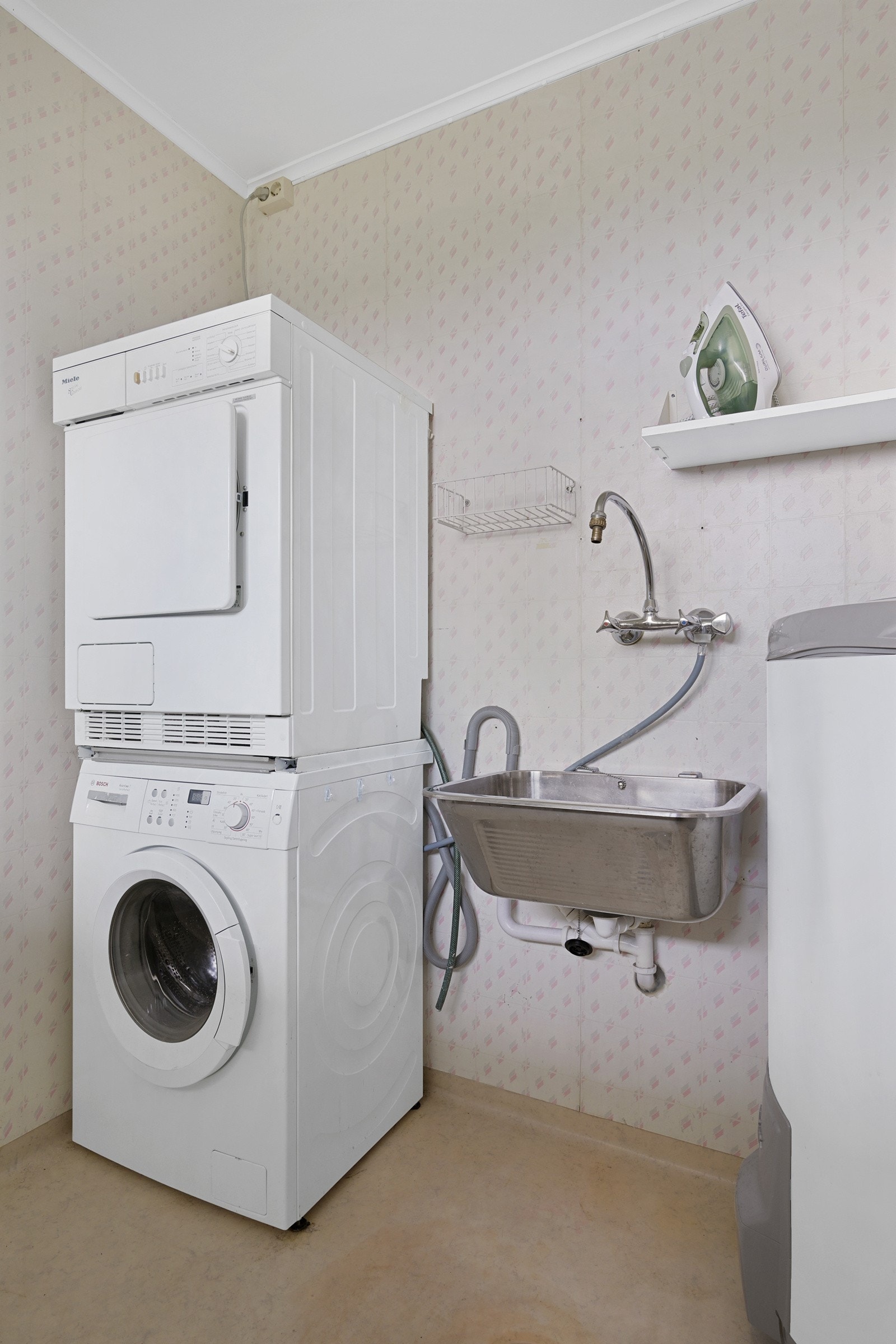 Praktisk vaskerom som er utstyrt med vinylbelegg på gulvet og tapetserte vegger. Rommet har en skyllekum av stål og opplegg for vaskemaskin Galleribilde