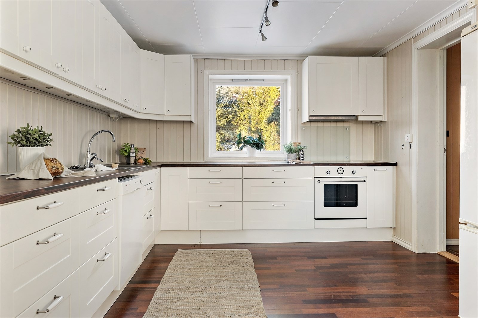 Romslig kjøkken med profilerte fronter og benkeplate i høytrykkslaminat. Det er integrert komfyr og platetopp fra 2024, frittstående kjøl/fryseskap og opplegg for oppvaskmaskin Galleribilde