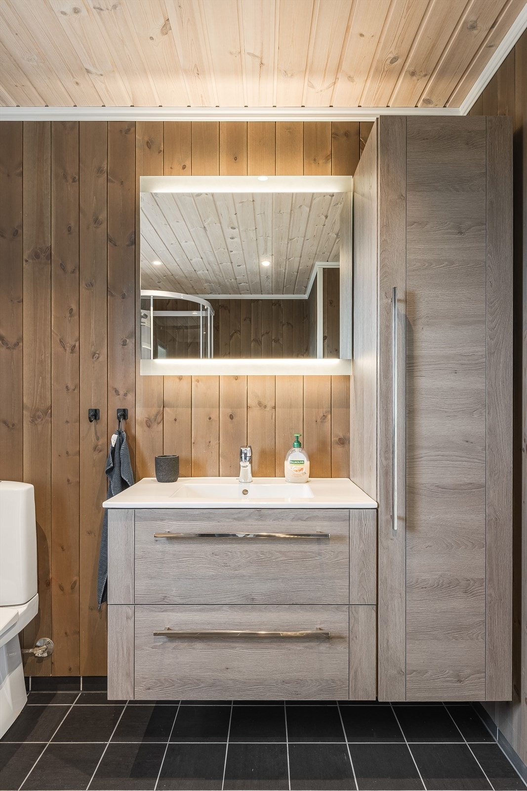 Pen baderomsinnredning med skuffer og heldekkende servant, speil med belysning og ekstra høyskap ved siden av. Galleribilde