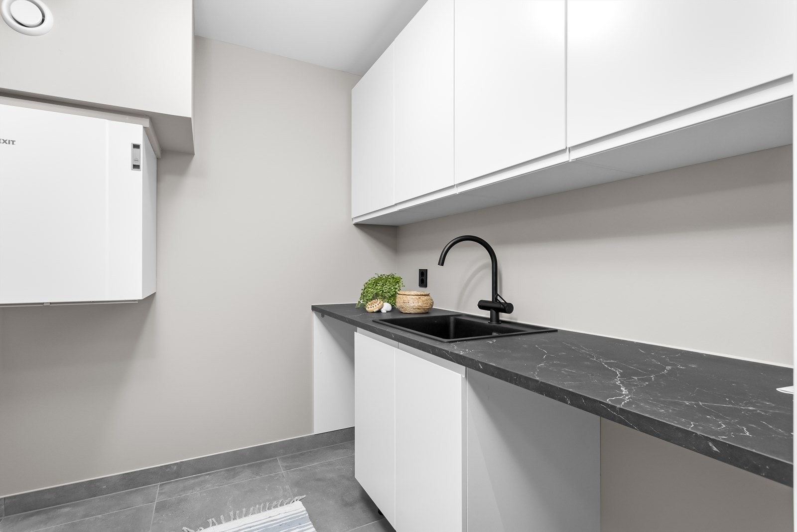 Praktisk vaskerom med oppbevaringsmuligheter i overskap, og en funksjonell benkeplate med nedfelt vask og avsatt plass til vaskemaskin og tørketrommel. Galleribilde