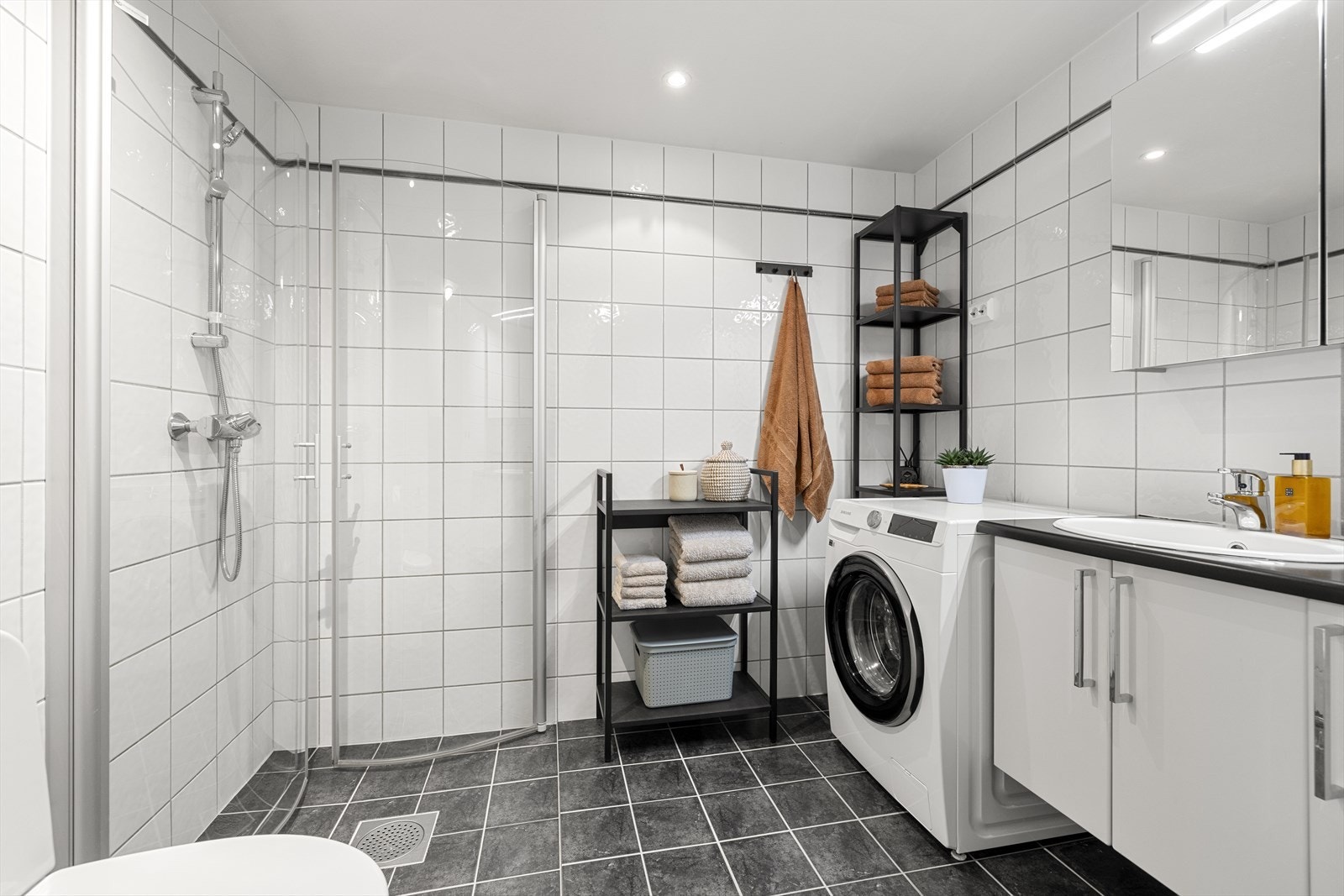Badet er innredet med hvite veggfliser og grå gulvfliser. Her er det toalett, dusjhjørne, servant, veggskap og et speil med belysning. Galleribilde