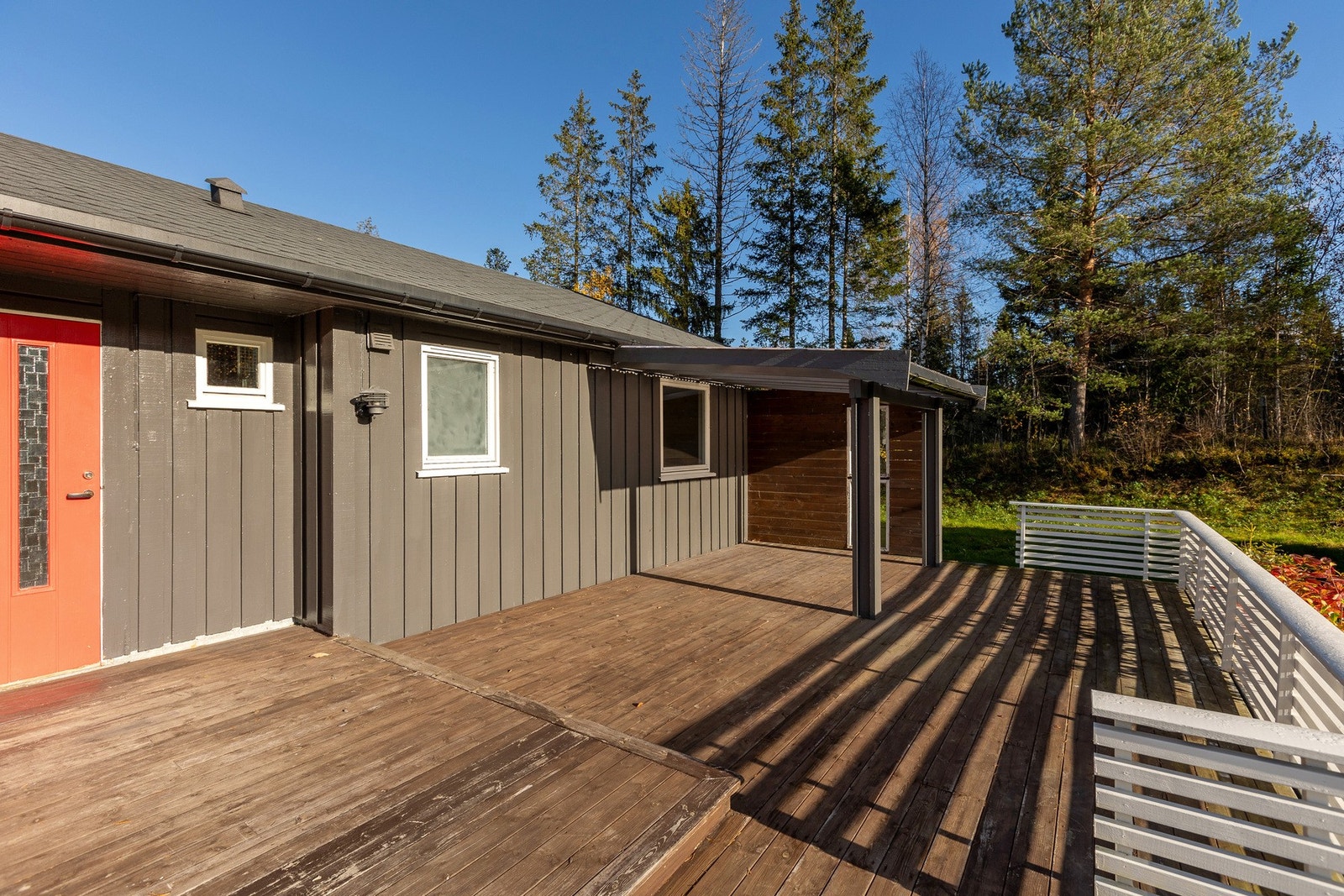 Terrasse med overbygg og trepaneler, omringet av natur. Vegger malt i grått med rødt inngangsparti. Galleribilde