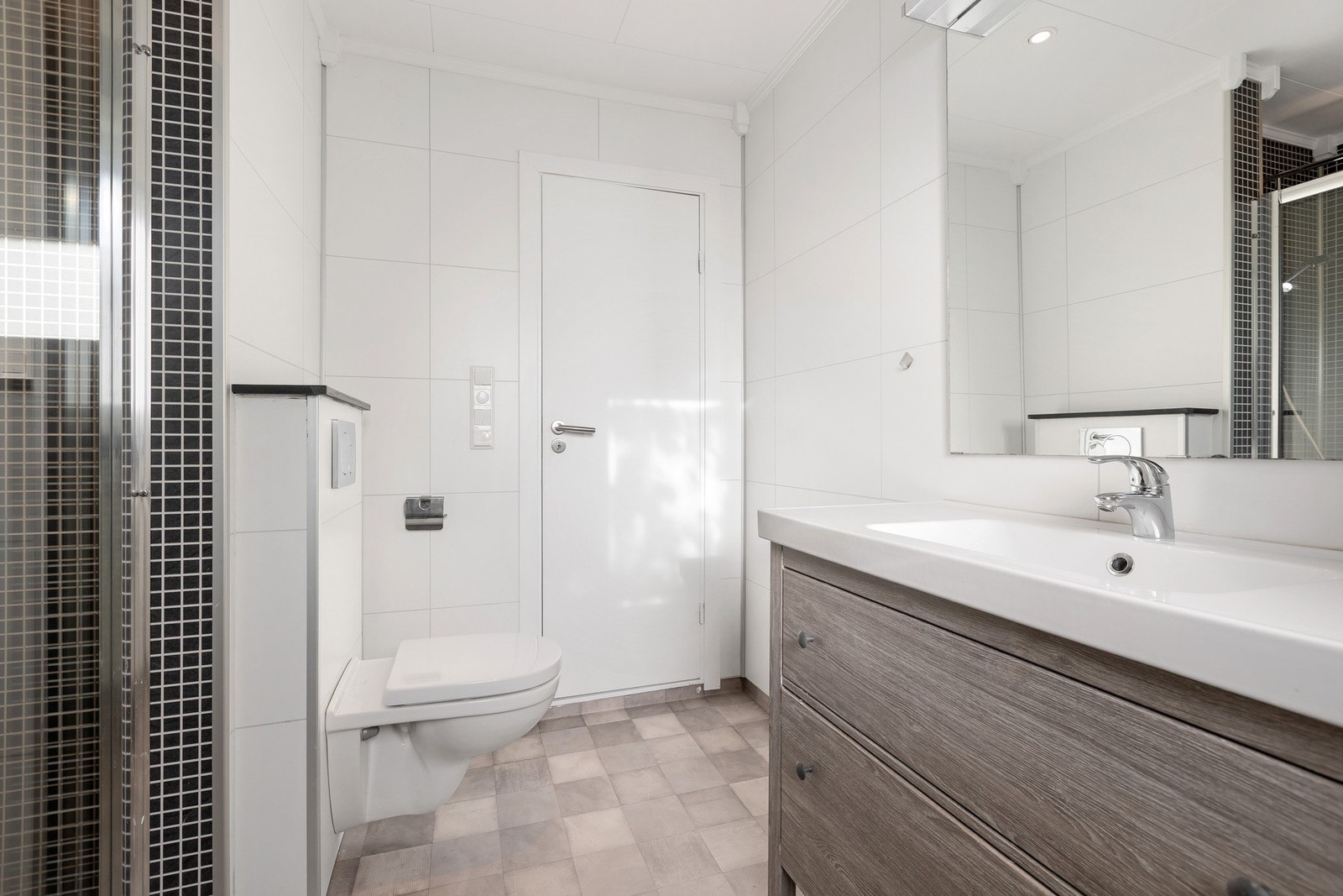 Lyst baderom med dusj, vegghengt toalett, servant og speil. Flislagt gulv og vegger. Galleribilde