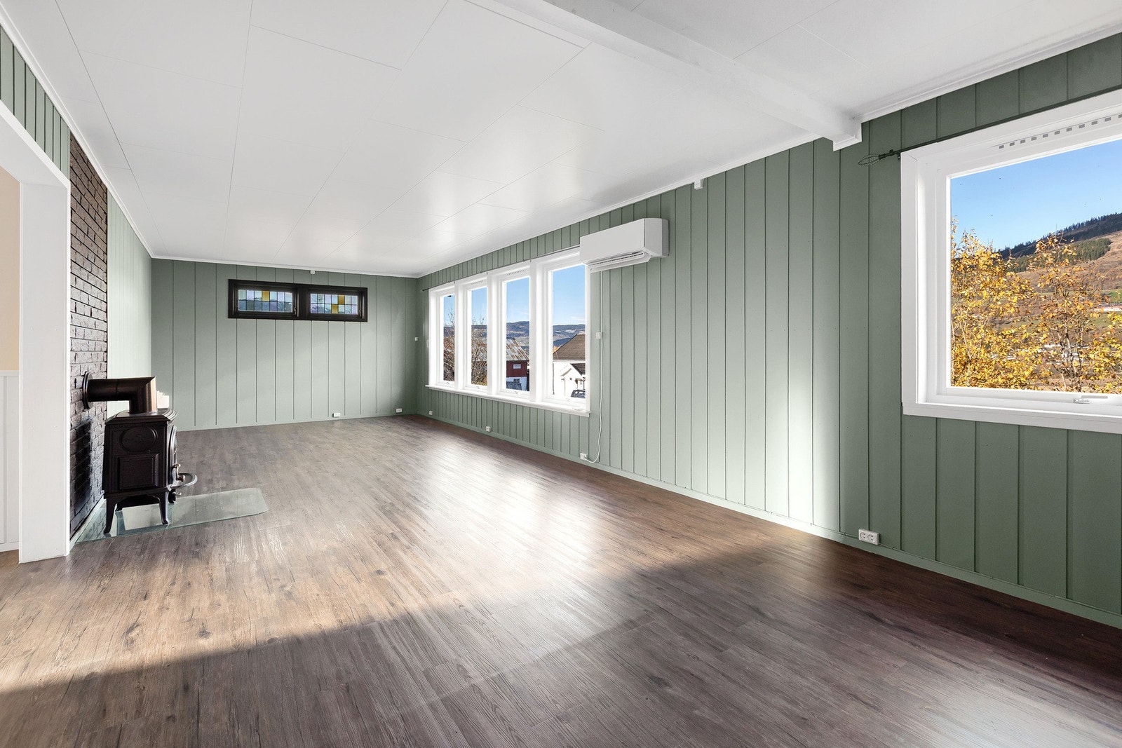 Stuen har grønne panelvegger, hvitt tak, store vinduer og laminatgulv. En vedovn står mot en mursteinsvegg. Galleribilde