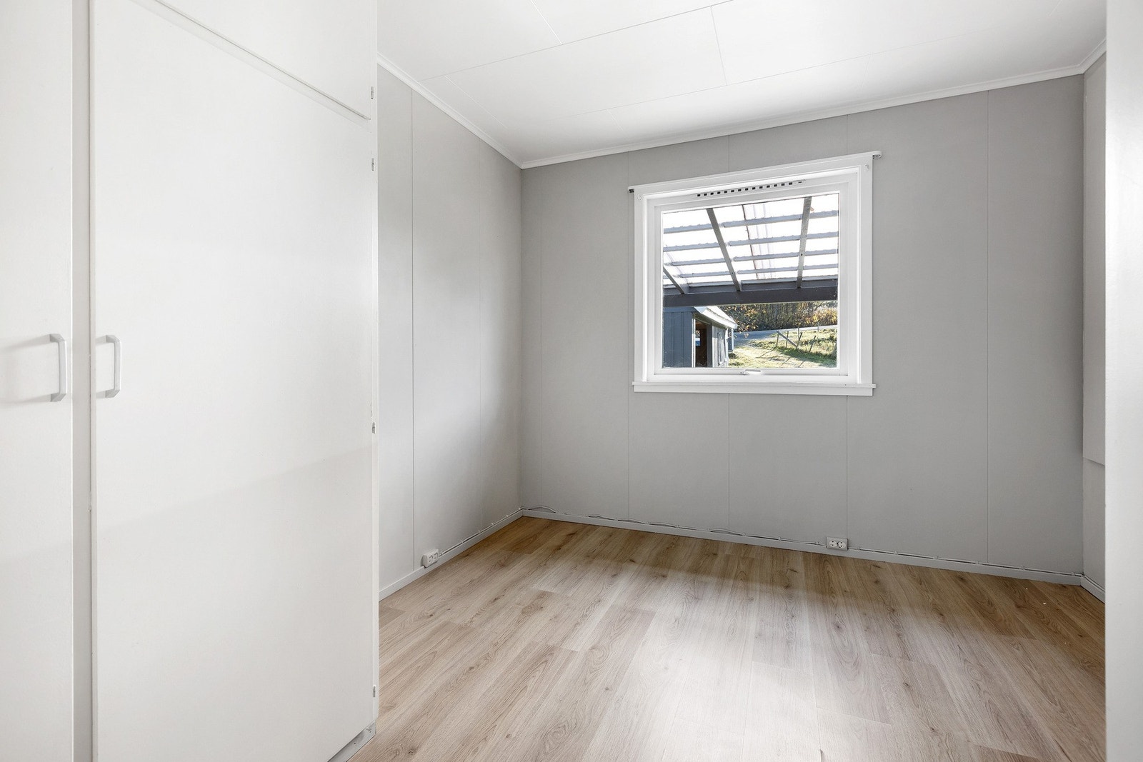 Rommet har grå vegger, hvitt tak og lys parkett. Vinduer gir utsikt mot hagen og overbygd uteområde. Galleribilde