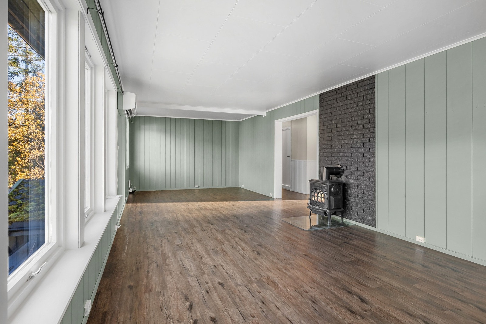 Rommet har store vinduer, peisovn, panelte vegger i grønt og grått, og mørkt laminatgulv. Galleribilde