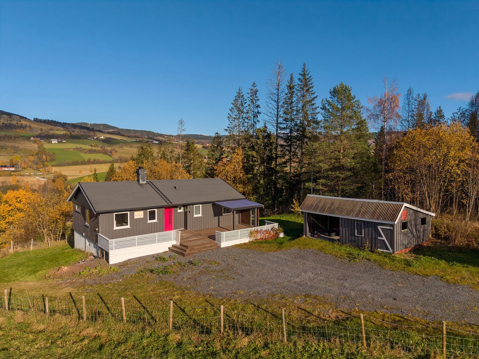 Enebolig med grå fasade, rødt inngangsparti, stor gruslagt gårdsplass og frittliggende garasje/ uthus. Omgitt av natur og landlig utsikt. Galleribilde