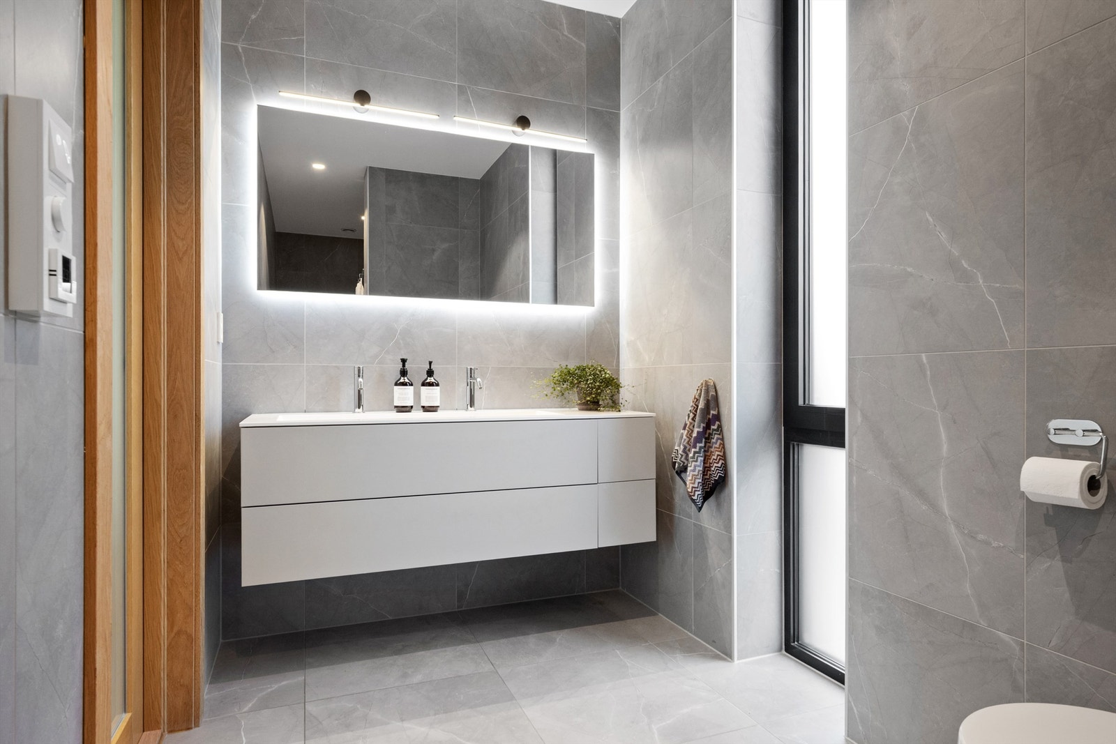 Badet er delikat utformet og av god størrelse og har lekre detaljer, flislagte vegger og gulv, servant med dobbel kran, badekar, dusjhjørne og vegghengt toalett. Galleribilde