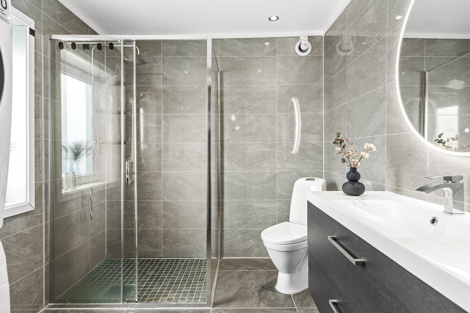 Moderne grå fliser på gulv, våtromsplater på vegg, toalett, servantinnredning, speil med integrert belysning og dusjnisje med glassdører. Galleribilde