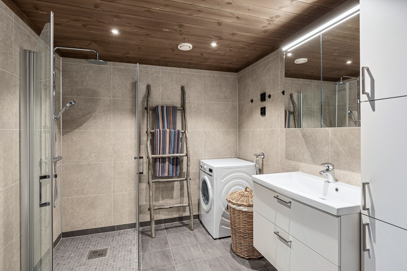 Badet er i lyse farger og har fliser på gulv, våtromsplater på vegg og panel i himling. Rommet er innredet med servant med underskap, dusjhjørne og vegghengt toalett. Galleribilde