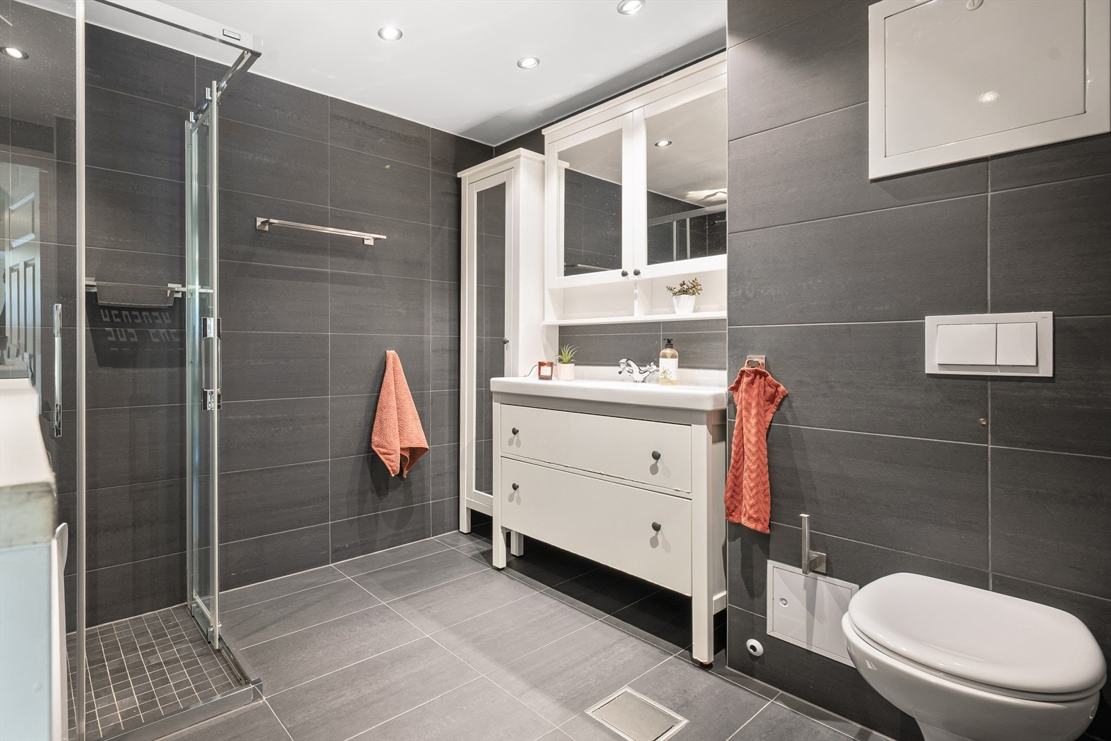 Pent flislagt baderom med tidløse og moderne flisvalg, vegghengt wc, helservant på baderomsinnredning og dusjhjørne med skyvedører i herdet glass. Galleribilde