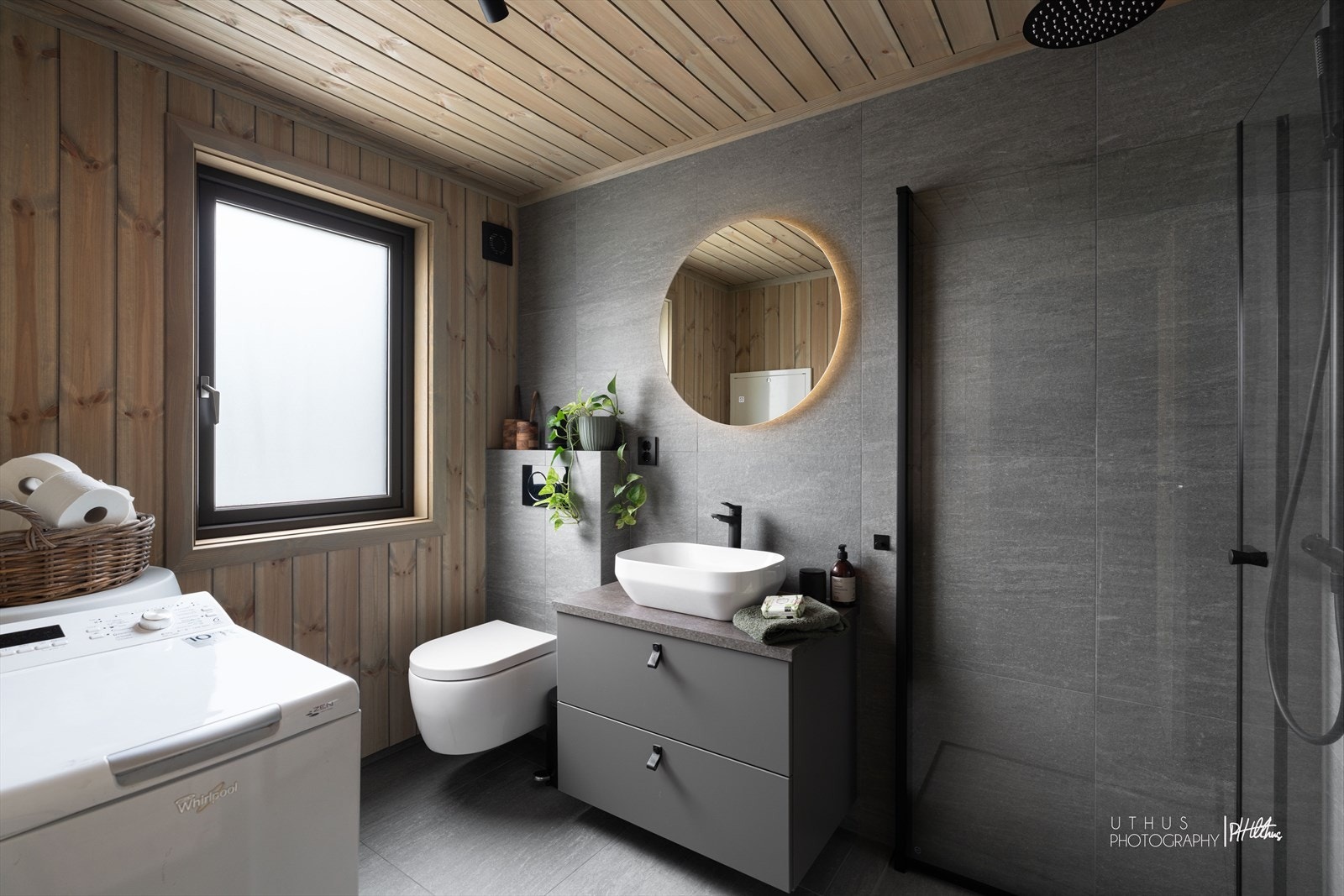 Stilrent bad med fliser, gulvvarme og en lun kontrastvegg i furu. Badet er utstyrt med dusjhjørne, servantskap, speil, vegghengt toalett og opplegg til vaskemaskin. Galleribilde