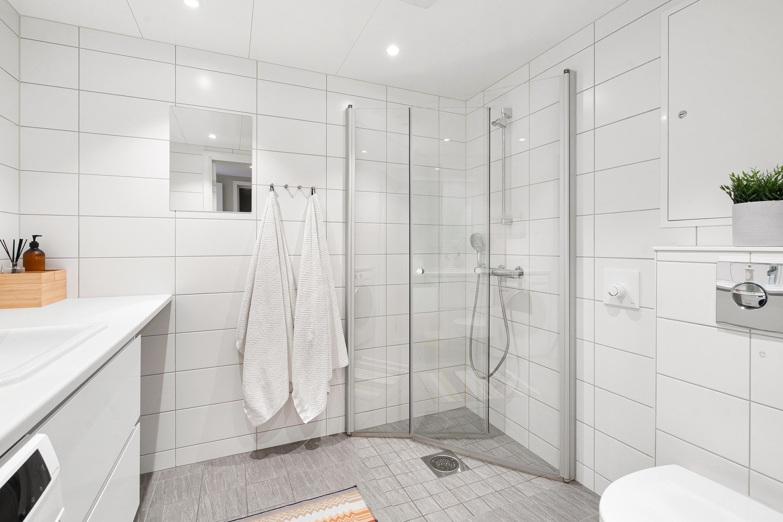 Badet er utstyrt med vegghengt benkeplate av laminat, nedfelt servant med ett-greps armatur, speil og lysarmatur med stikkontakt over servant samt vegghengt. Videre er det dusjhjørne med innfellbare glassdører med dusjarmatur tilkoblet hånddusj. Galleribilde