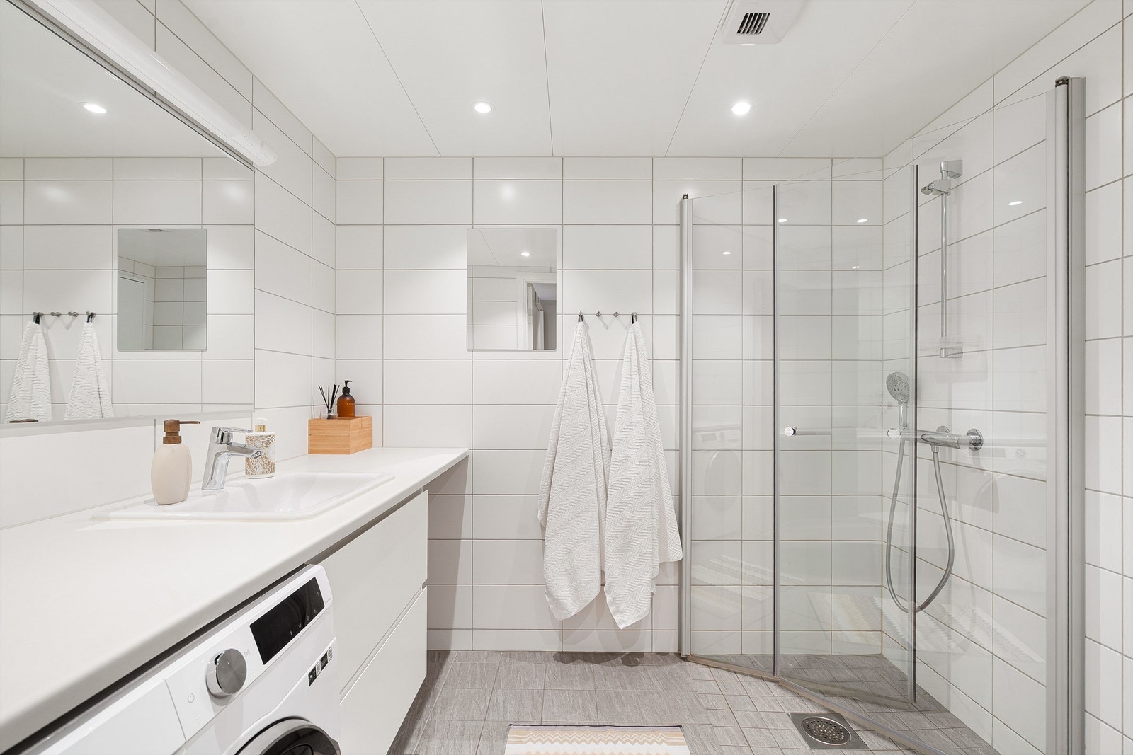 Romslig baderom fra byggeår (2022) med opplegg for vaskemaskin, flislagt gulv med gulvvarme, flislagte vegger og hvit lakkerte metallplater i himling med downlights. Galleribilde