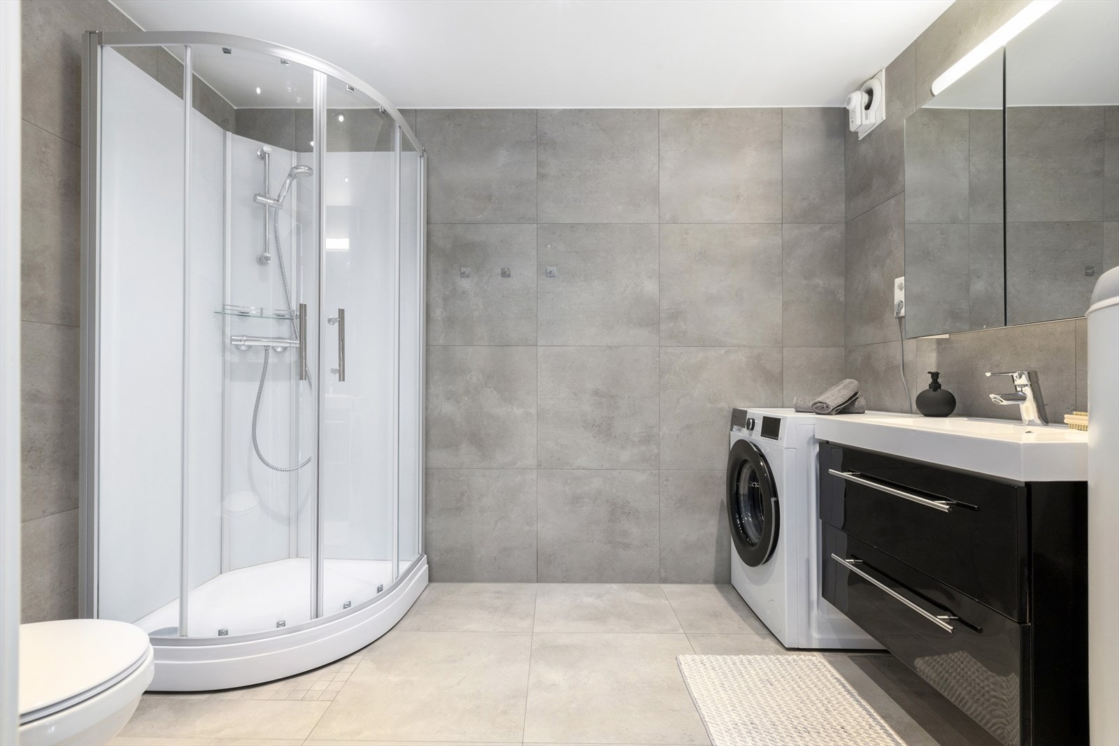 Moderne flislagt bad med varmekabler i gulv og downlights i tak. Badet er pent innredet med sort baderomsinnredning med heldekkende nedfelt servant, speil med belysning, vegghengt toalett og dusjkabinett. Galleribilde