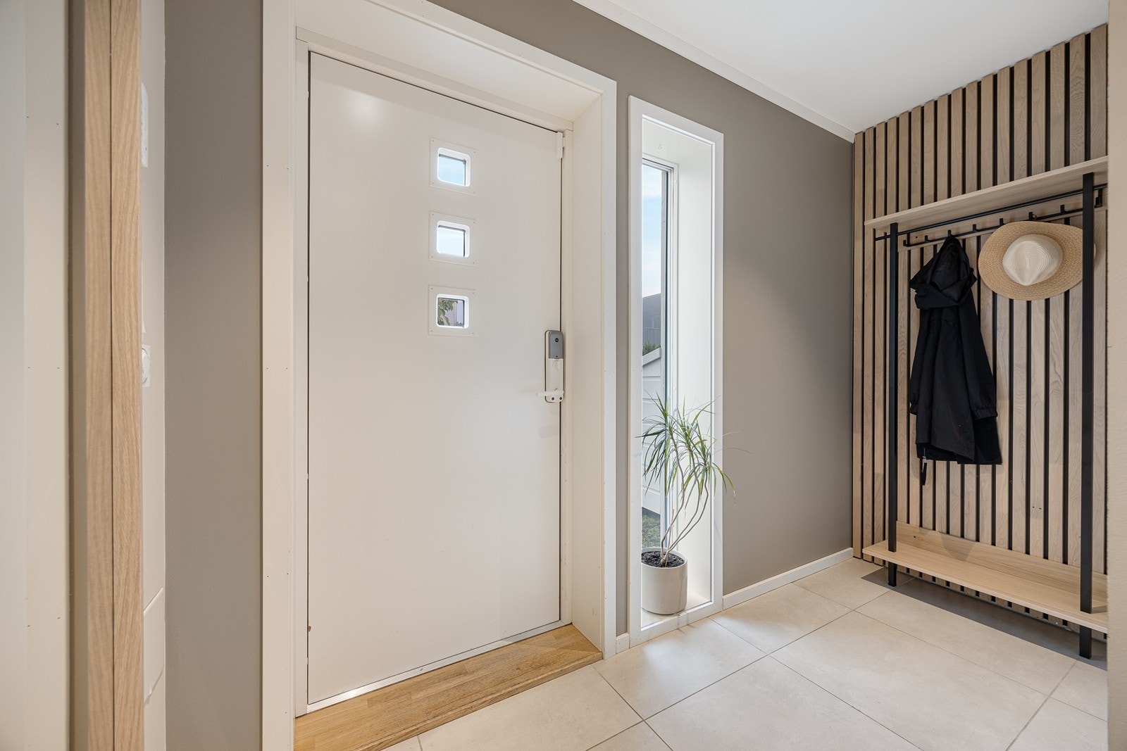 Inne venter en moderne entré med store, beige gulvfliser, gråmalte vegger og en fondvegg med trespiler Galleribilde