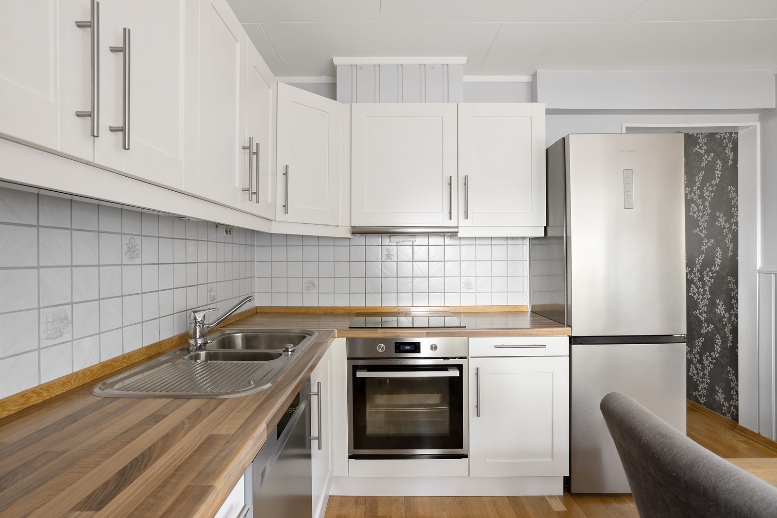 Innredningen har profilerte fronter, benkeplater i eikemønstret laminat, benkebelysning og hvite fliser med stedvis dekor på veggene bak. Galleribilde