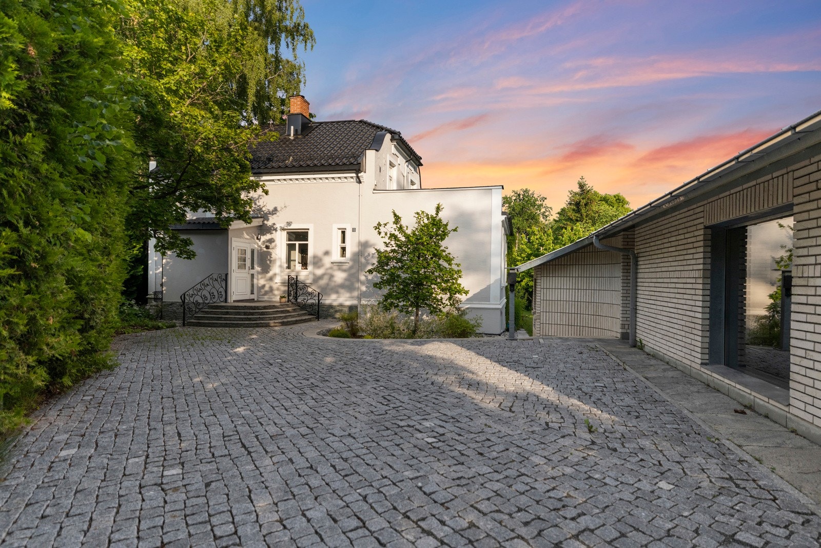 Brosteinsbelagt gårdsplass med frittstående dobbelgarasje, skrivestue og elbillader både i garasje og på ladestolpe. Galleribilde