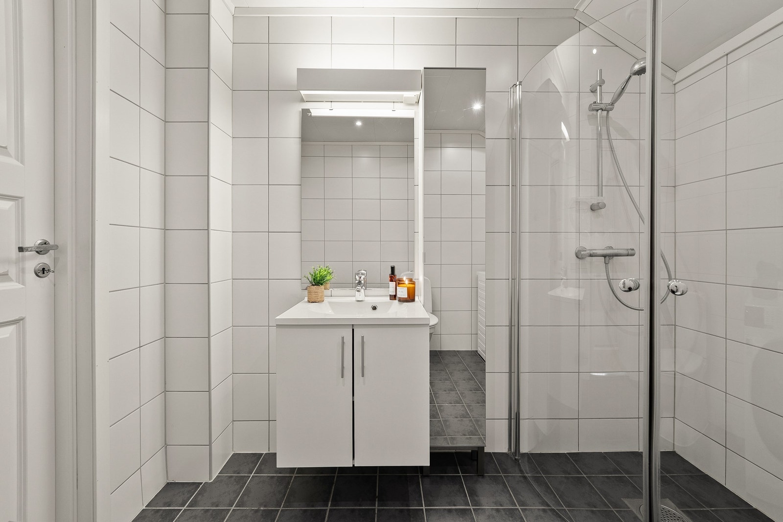 Det er vegghengt servantskap med ovenpåliggende servant, speil og belysning med stikkontakt over servant, vegghengt høyskap, gulvstående toalett, opplegg for vaskemaskin, og dusj på gulv med innfellbare dører av glass og metall. Galleribilde