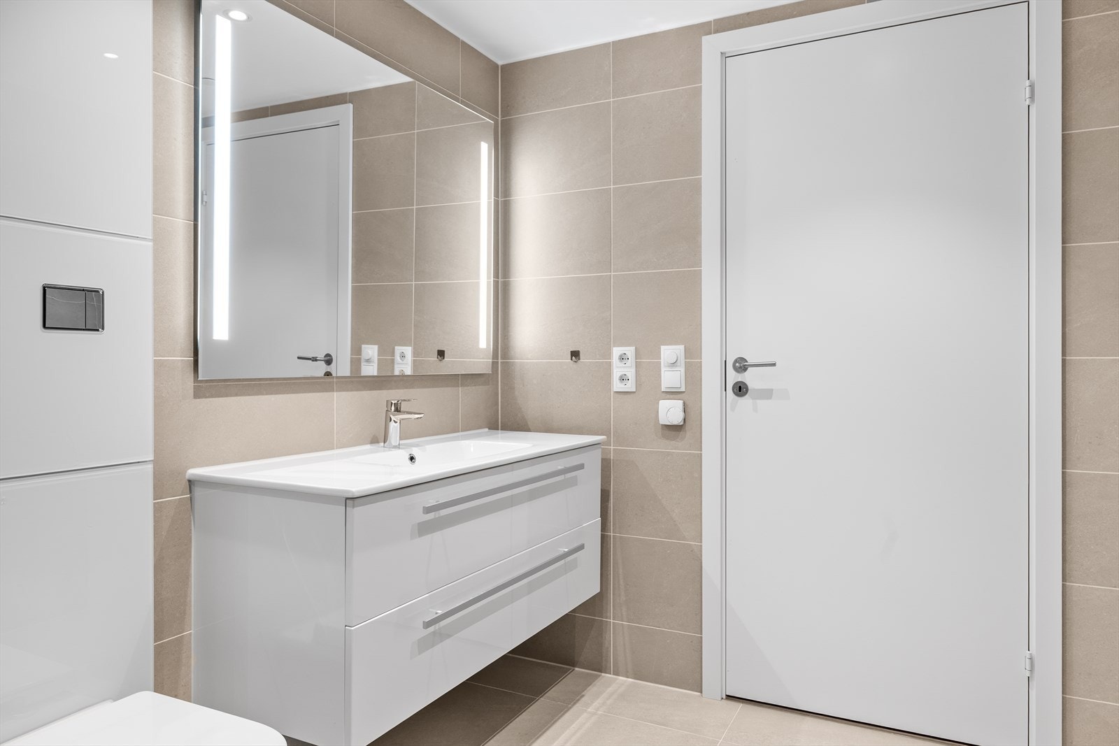 Delikat bad/vaskerom med flotte fliser på gulv og vegg. Bader er innredet med vegghengt toalett, dusj på vegg adskilt med svingbare dusjdører. Nedfelt servant i baderomsinnredning og opplegg til vaskemaskin. Galleribilde