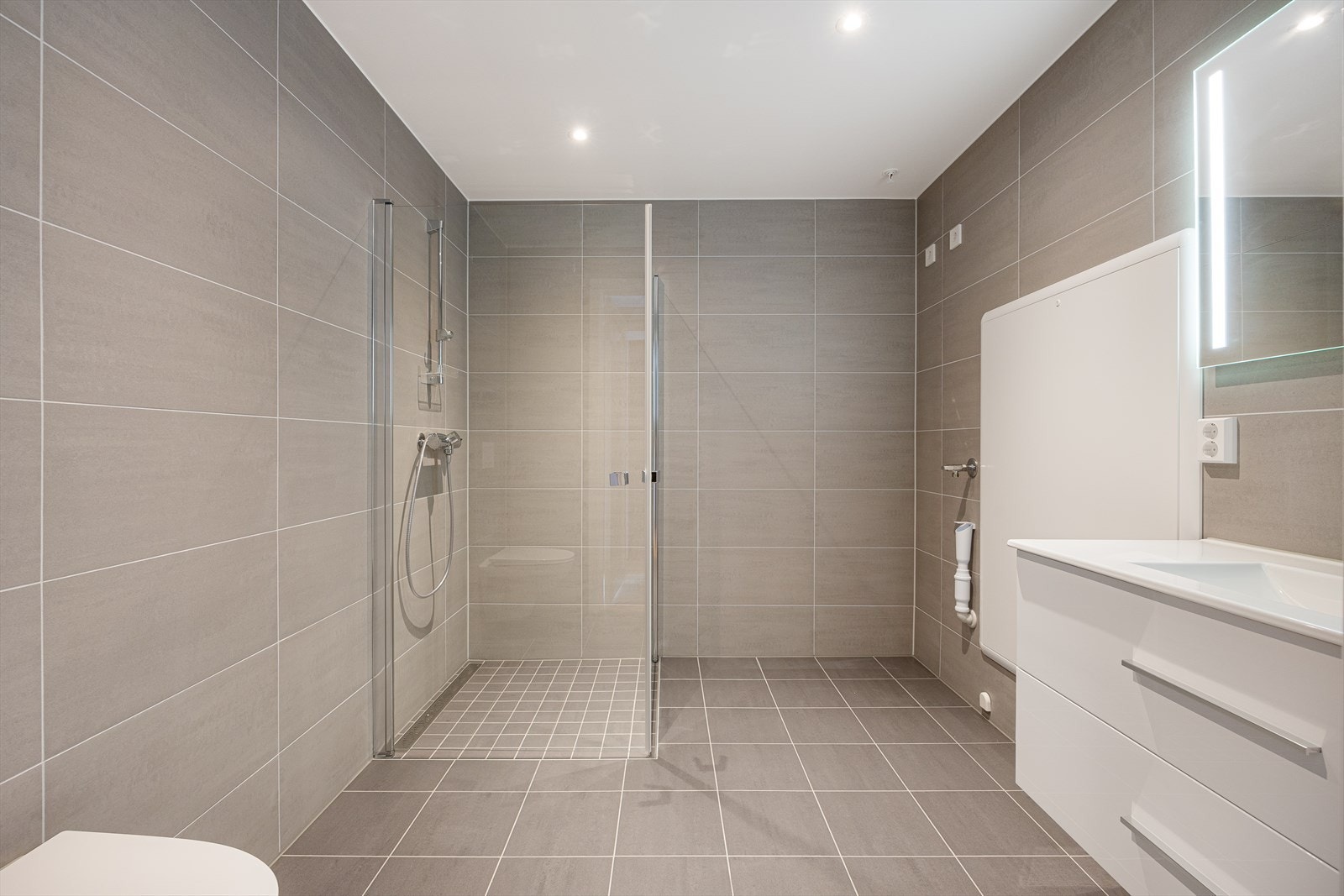 Badet er delikat utformet med grå fliser på gulv og vegger, gulvvarme og downlights Galleribilde