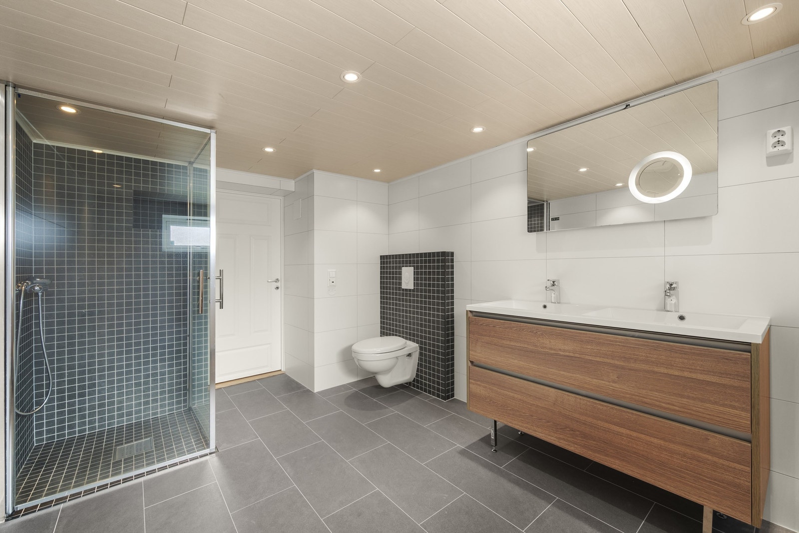 Moderne bad fra 2025 med fliser på gulv med varme og vegger. Nedfelt himling med downlights, dobbel servant med slette fronter, vegghengt toalett og dusjhjørne med innfellbare glassdører Galleribilde