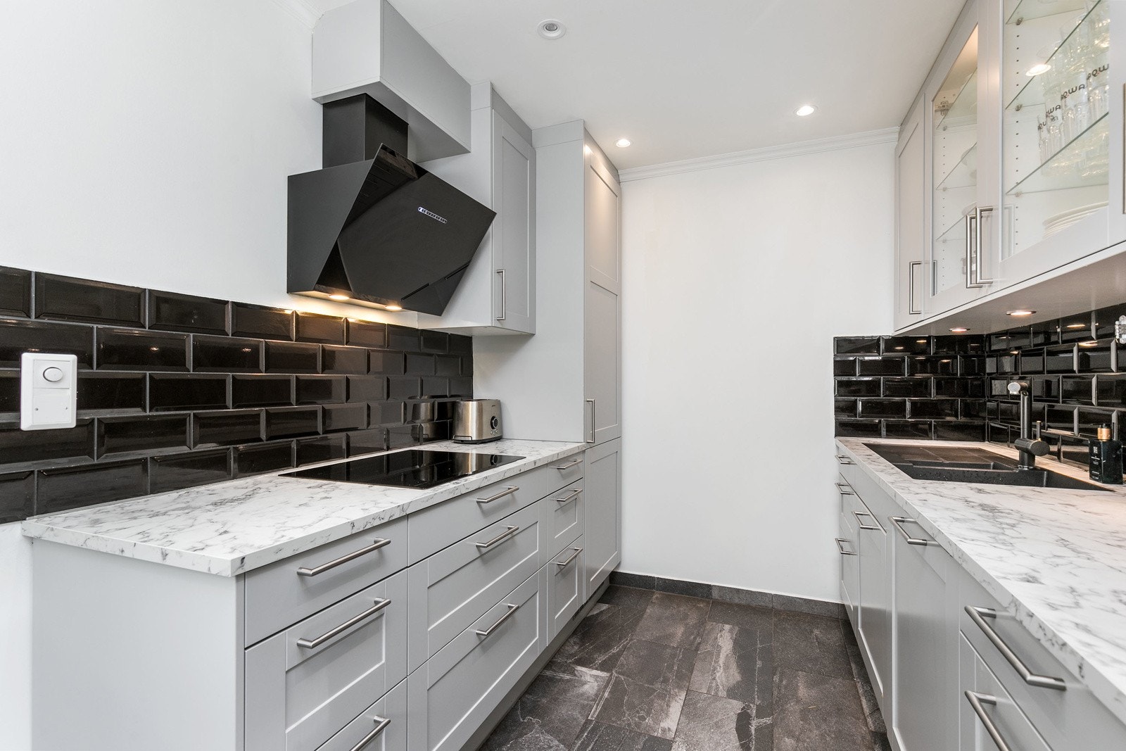 Innredningen består av profilerte fronter, enkelte glassfronter og laminat benkeplate med nedfelt kum. Mørke fliser på vegg over kjøkkenbenk og belysning under overskap. Galleribilde