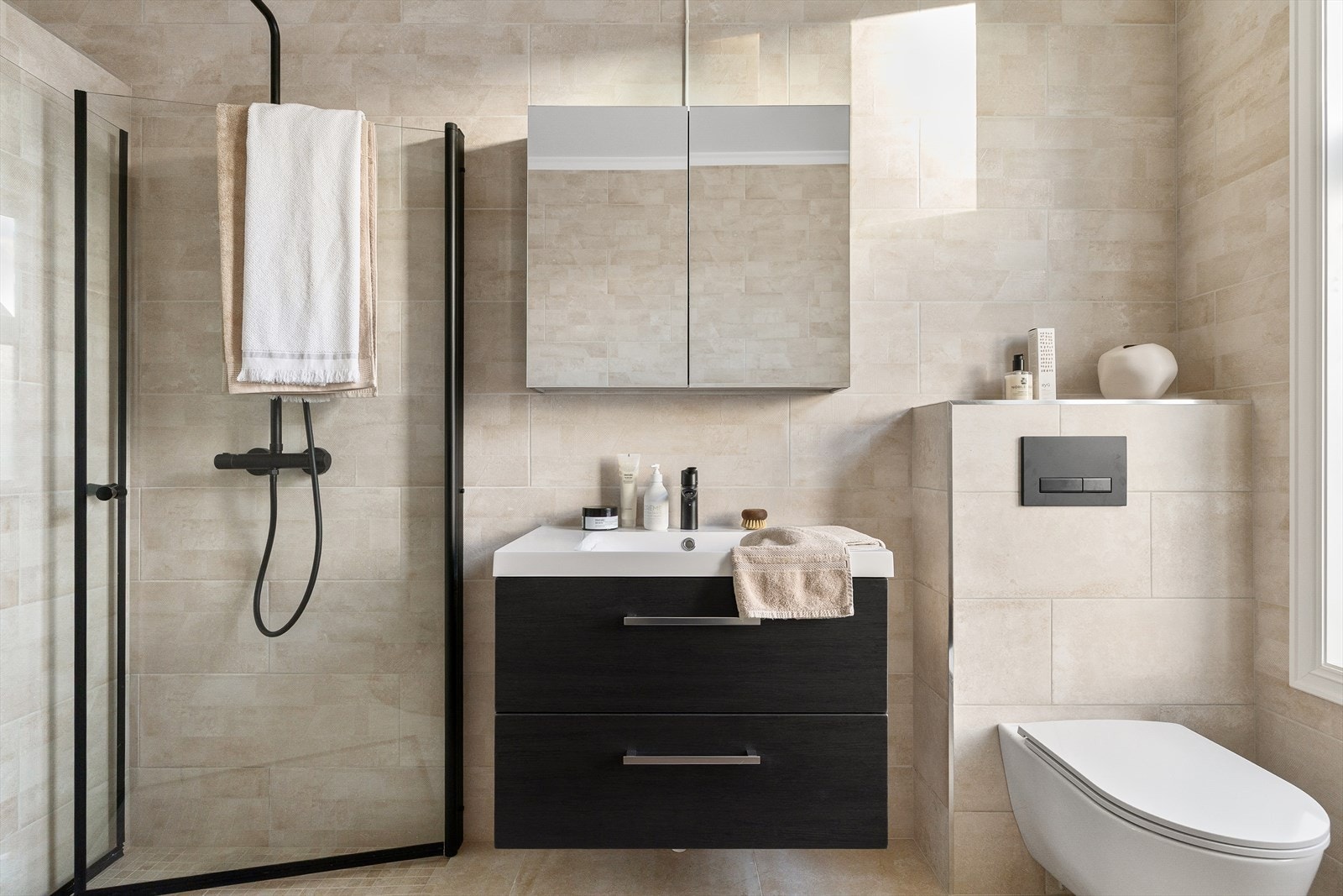 Badet er utstyrt med veggmontert toalett, moderne dusjhjørne med vegg og dør i herdet glass, samt servant med tilhørende skap, speil og integrert belysning. Galleribilde