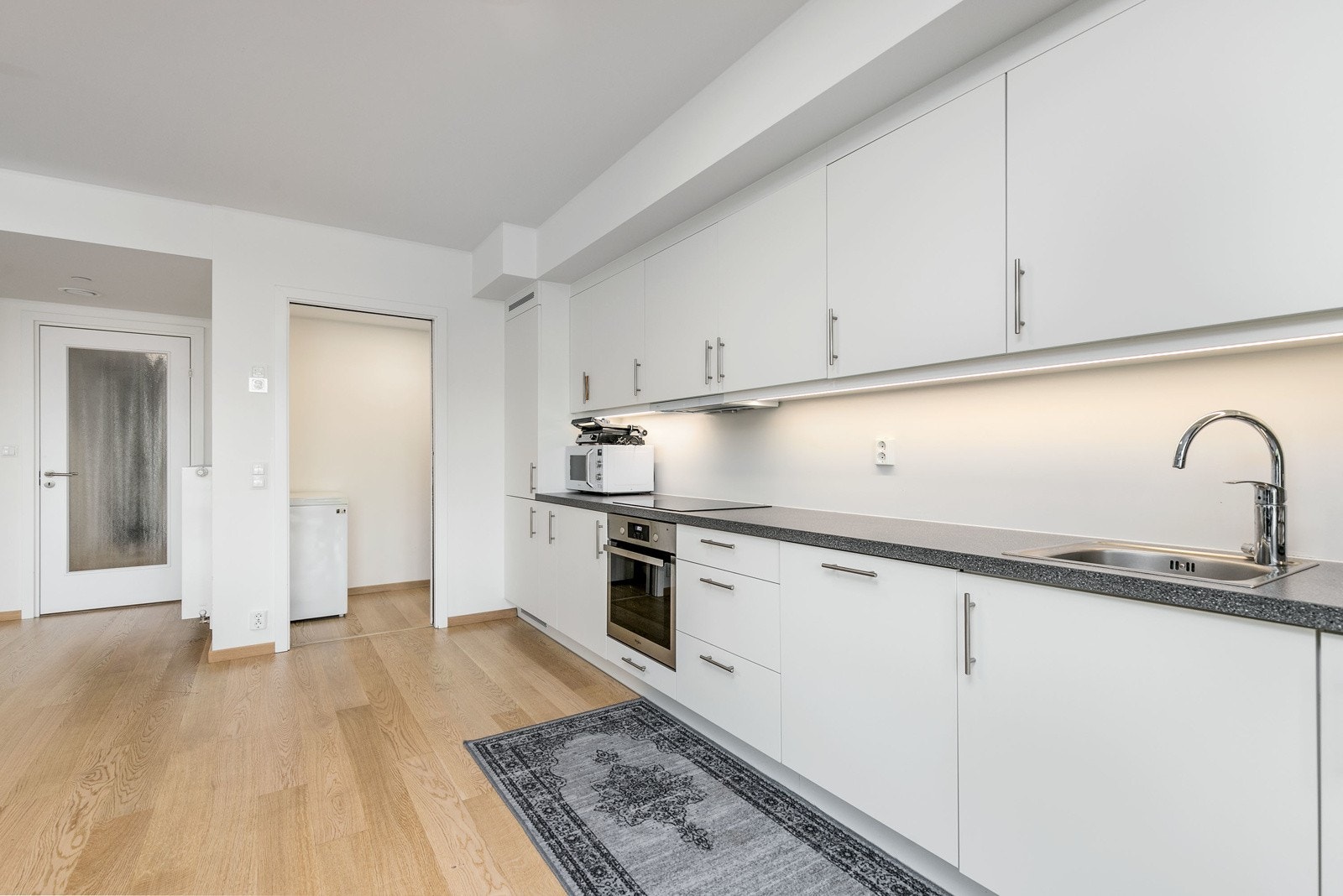 Nedfelt vaskekum i rustfritt stål med ett-greps blandebatteri. Det er satt inn lyslist mellom over- og underskap. Av utstyr medfølger integrerte hvitevarer som komfyr, induksjonsplatetopp, oppvaskmaskin, samt kombinert kjøle/- og fryseskap. Galleribilde