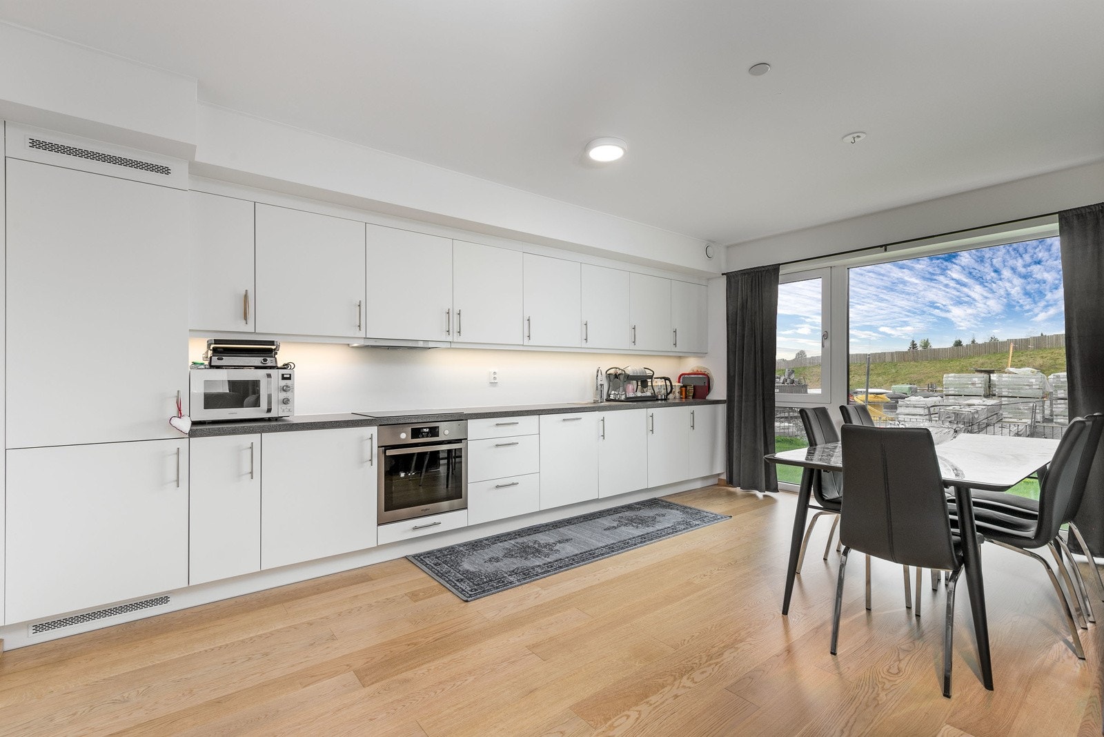 Stilrent kjøkken fra 2022 i åpen løsning mot stuen. Innredningen består av hvite slette fronter med foring opp til tak og laminert benkeplate i mørk utførelse. Galleribilde