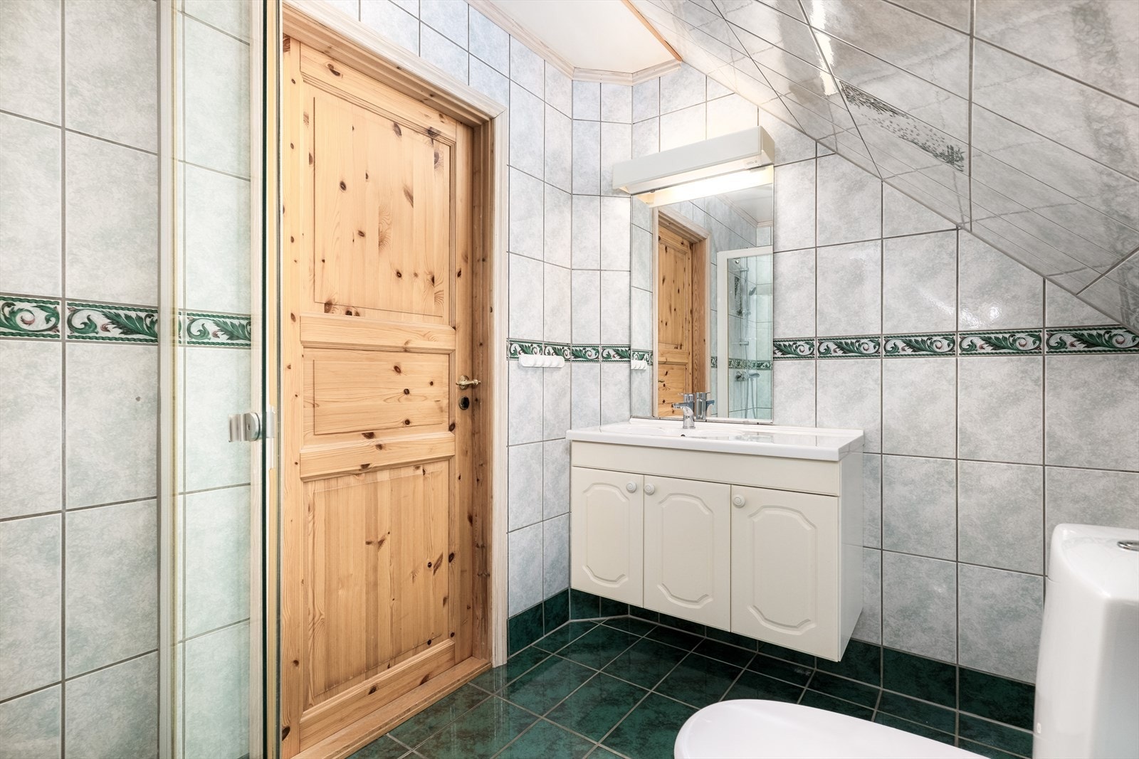 Flislagt bad med varmekabler i gulv. Badet er innredet med toalett, dusjhjørne med glassvegger og servant innredning med skap og speil. Galleribilde